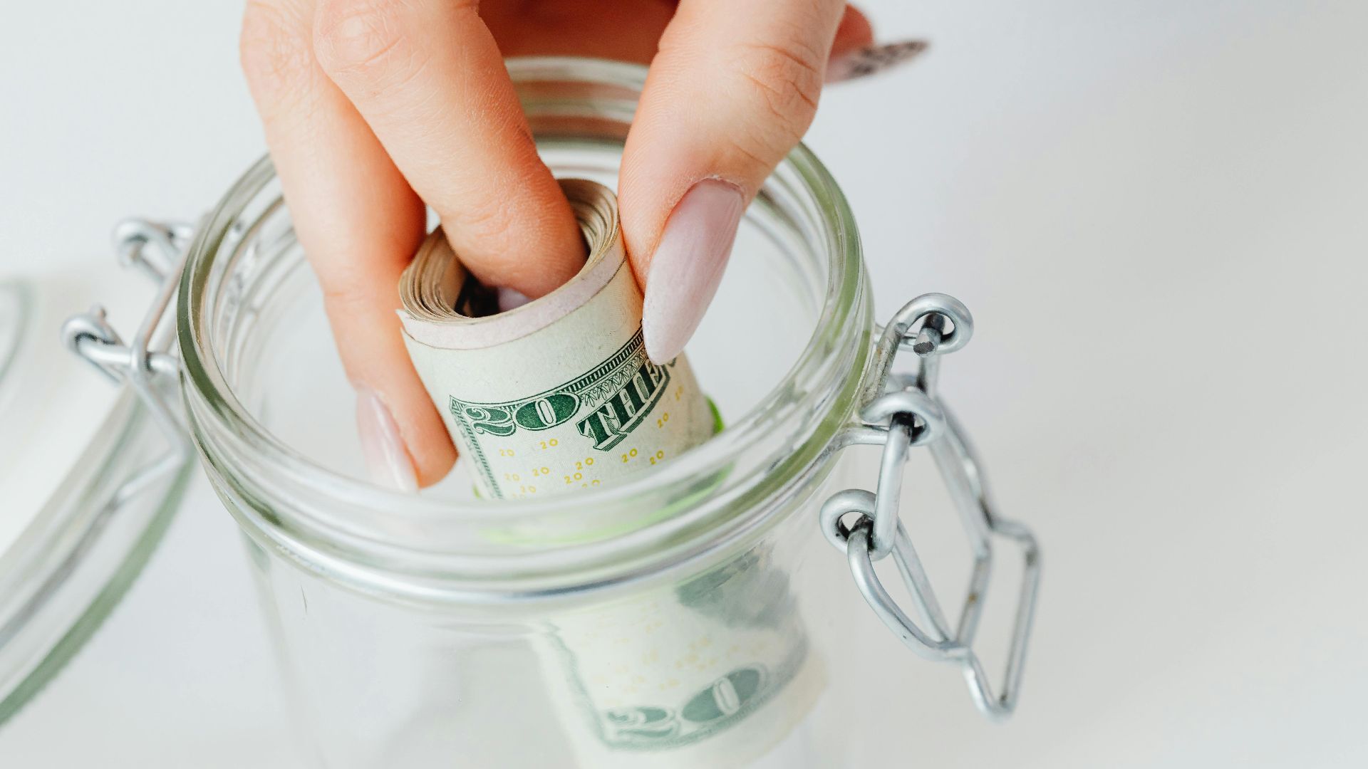 A close-up of a hand placing rolled dollars into a glass jar, symbolizing savings.
