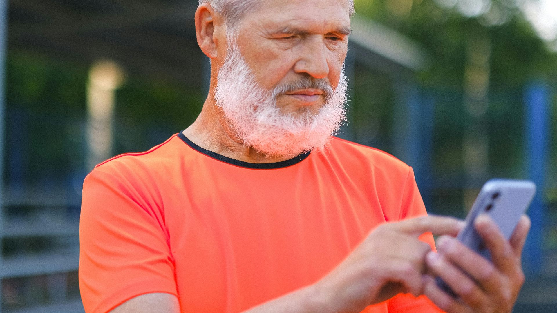 Crop elderly bearded male with gray hair browsing modern mobile phone while spending time in park