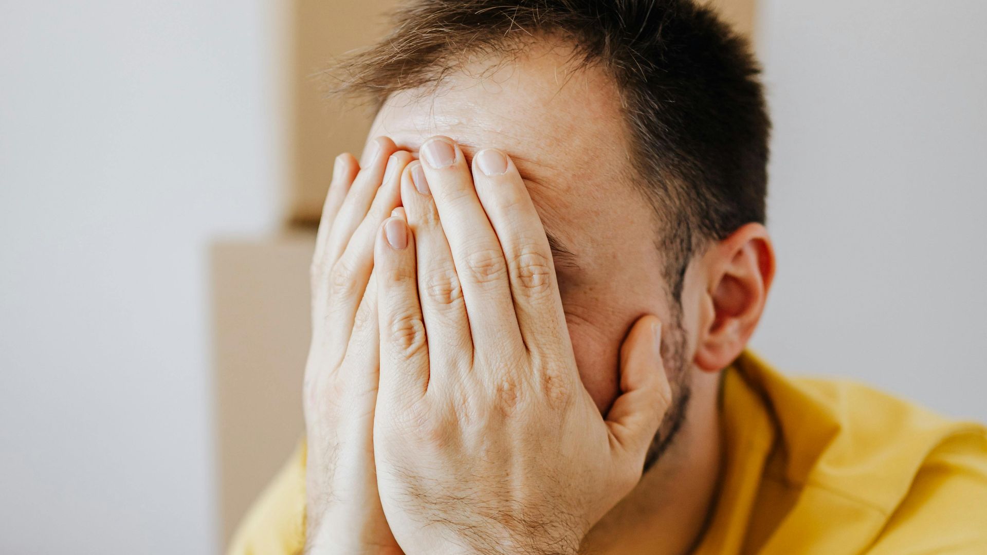 A stressed man sits with his hands covering his face amid moving boxes, depicting moving stress.