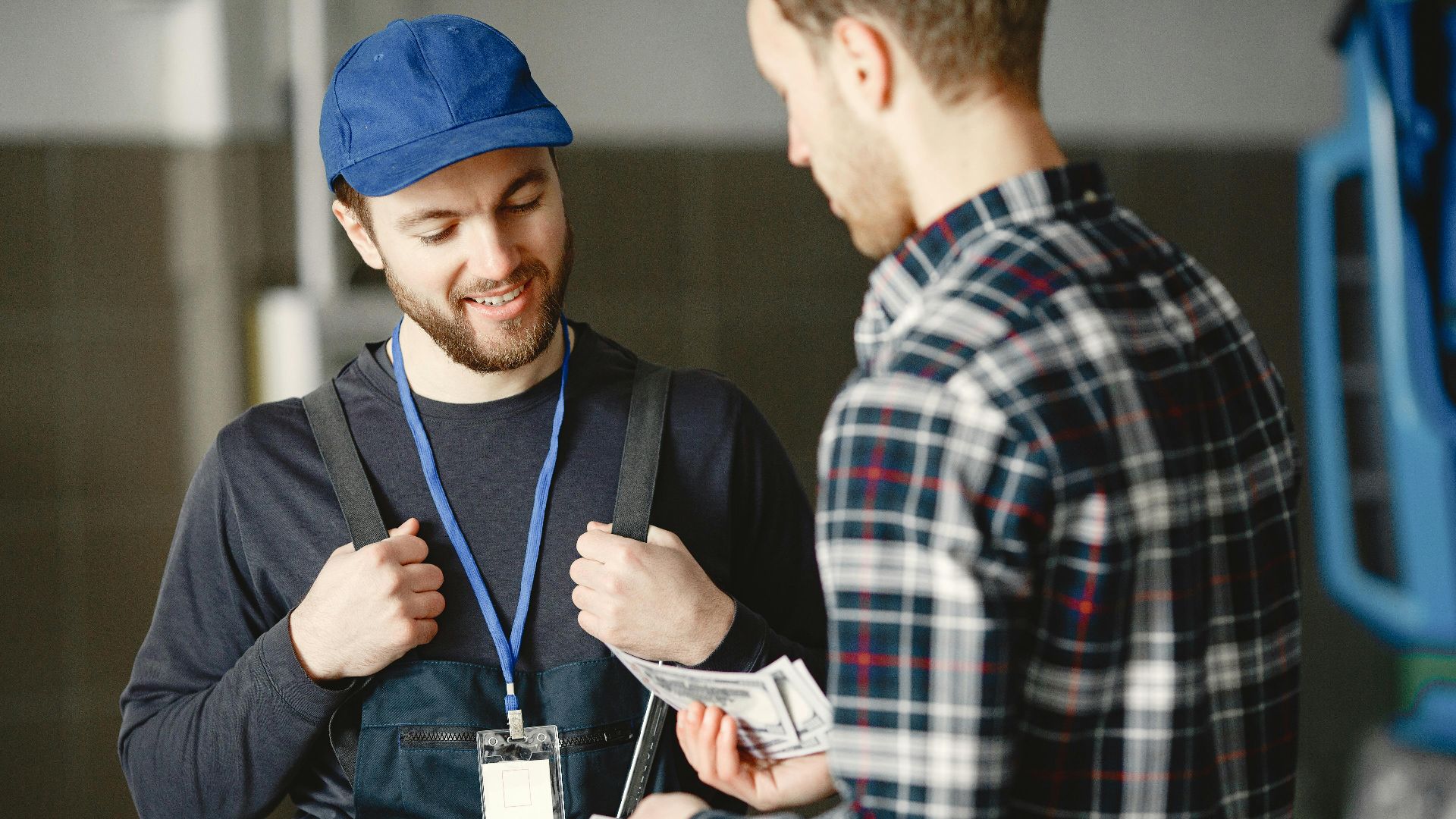 A mechanic in a blue cap receives cash payment from a customer in a plaid shirt, smiling.