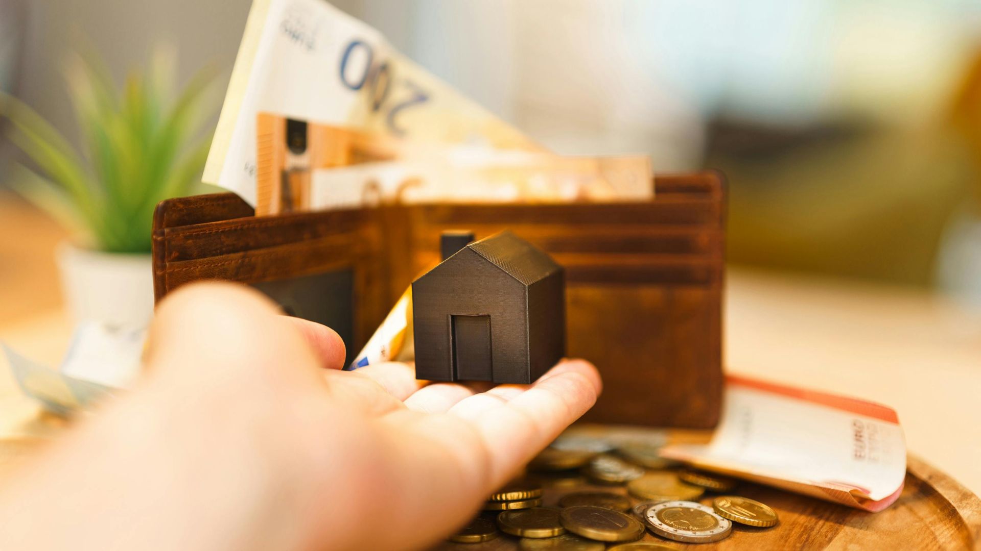 A hand holding a small house model with euro notes and coins nearby, illustrating real estate investment and finance.