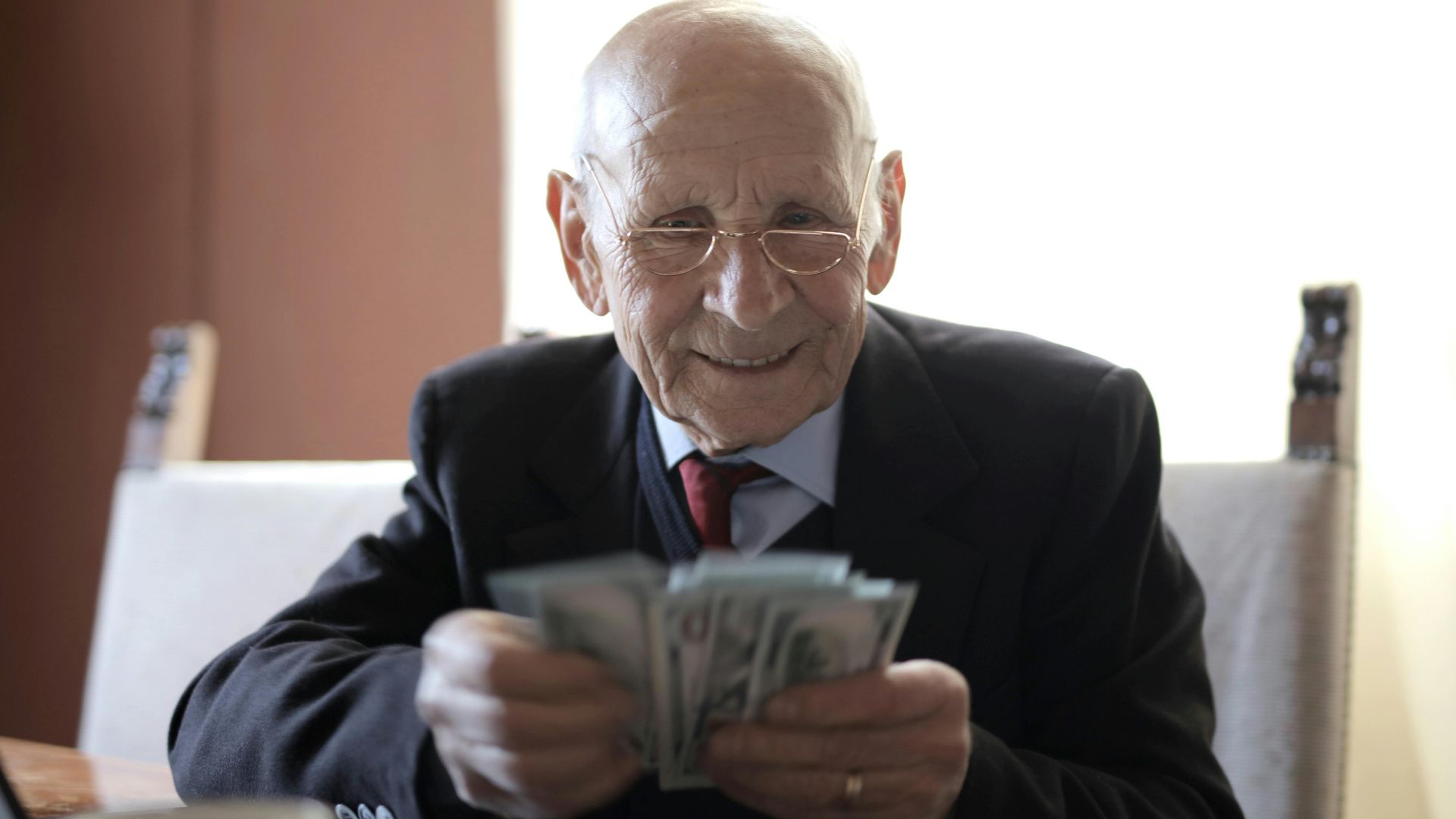 Positive senior businessman in formal suit and eyeglasses counting money bills while sitting at wooden table with cup of beverage and near opened laptop