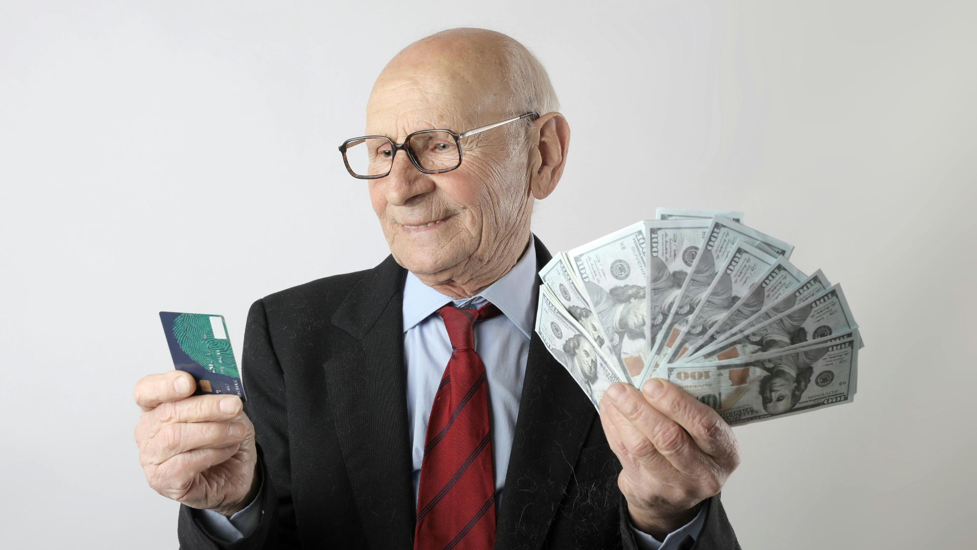 Elderly man in a suit holding a credit card and US dollar bills, representing finance and wealth.