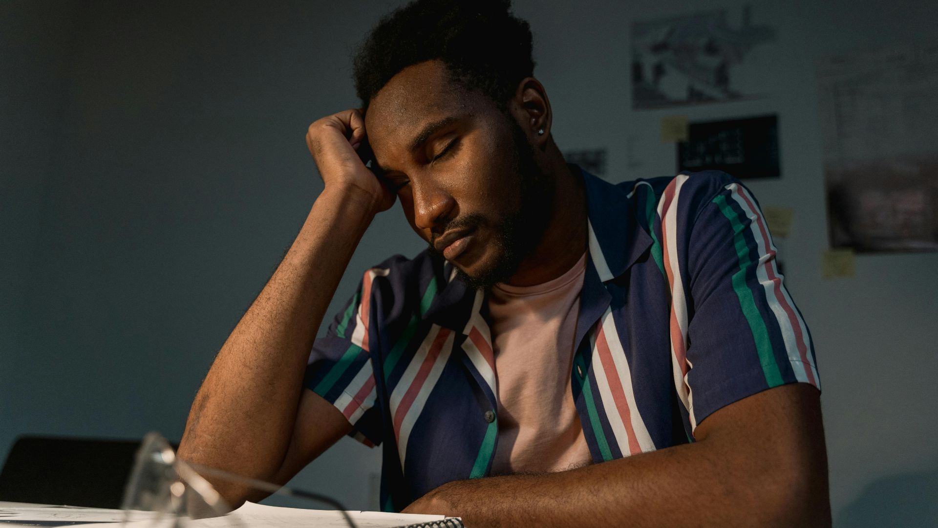 Man wearing a colorful striped shirt rests his head thoughtfully while sitting indoors.