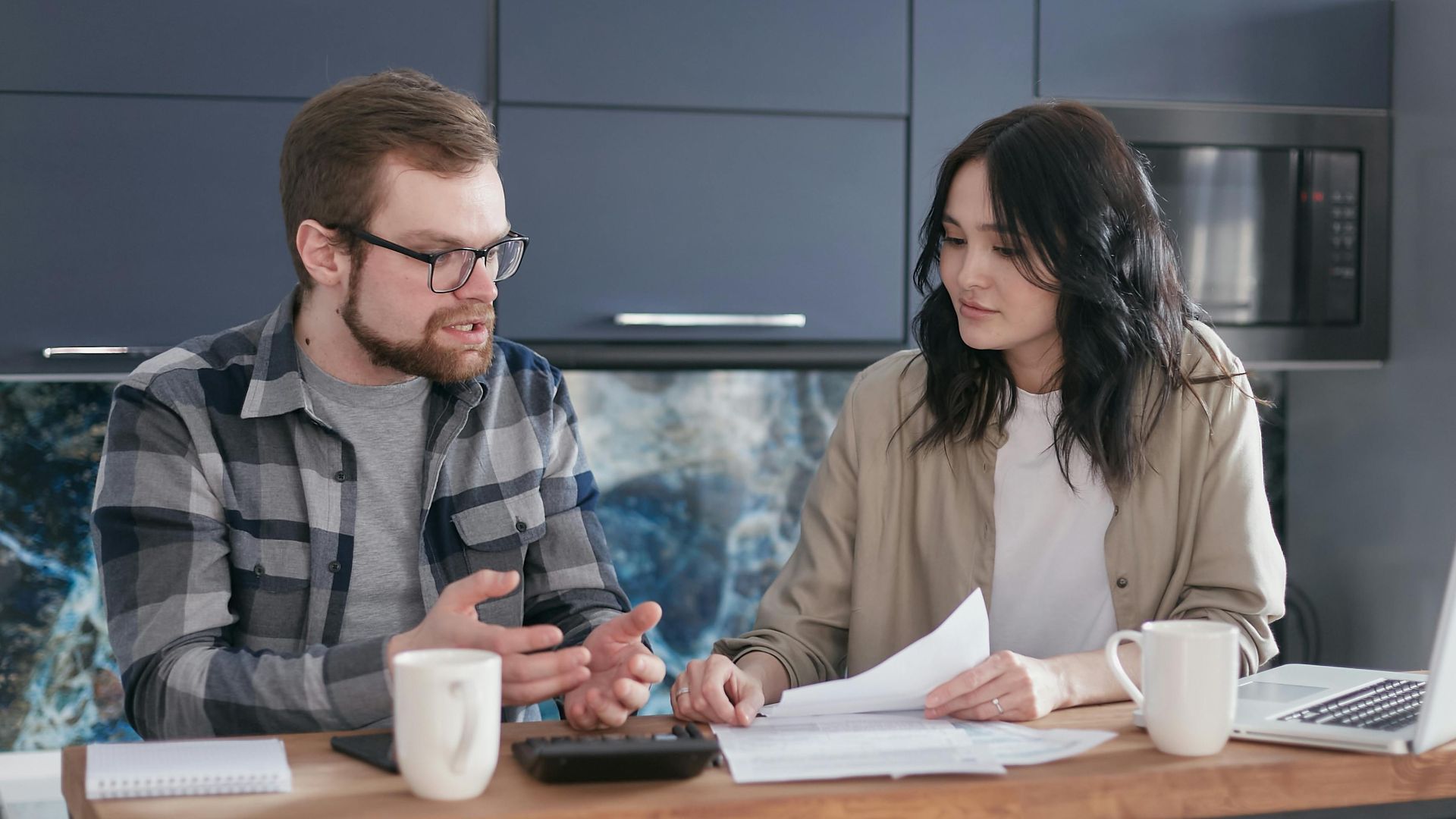 A couple engaged in a financial discussion, sitting at a kitchen table with papers and a laptop.