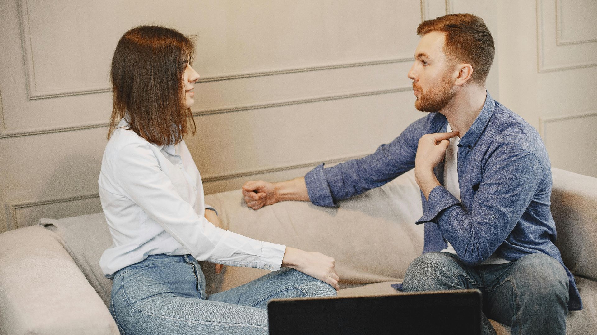 A young couple engaged in a conversation while sitting on a beige couch indoors.