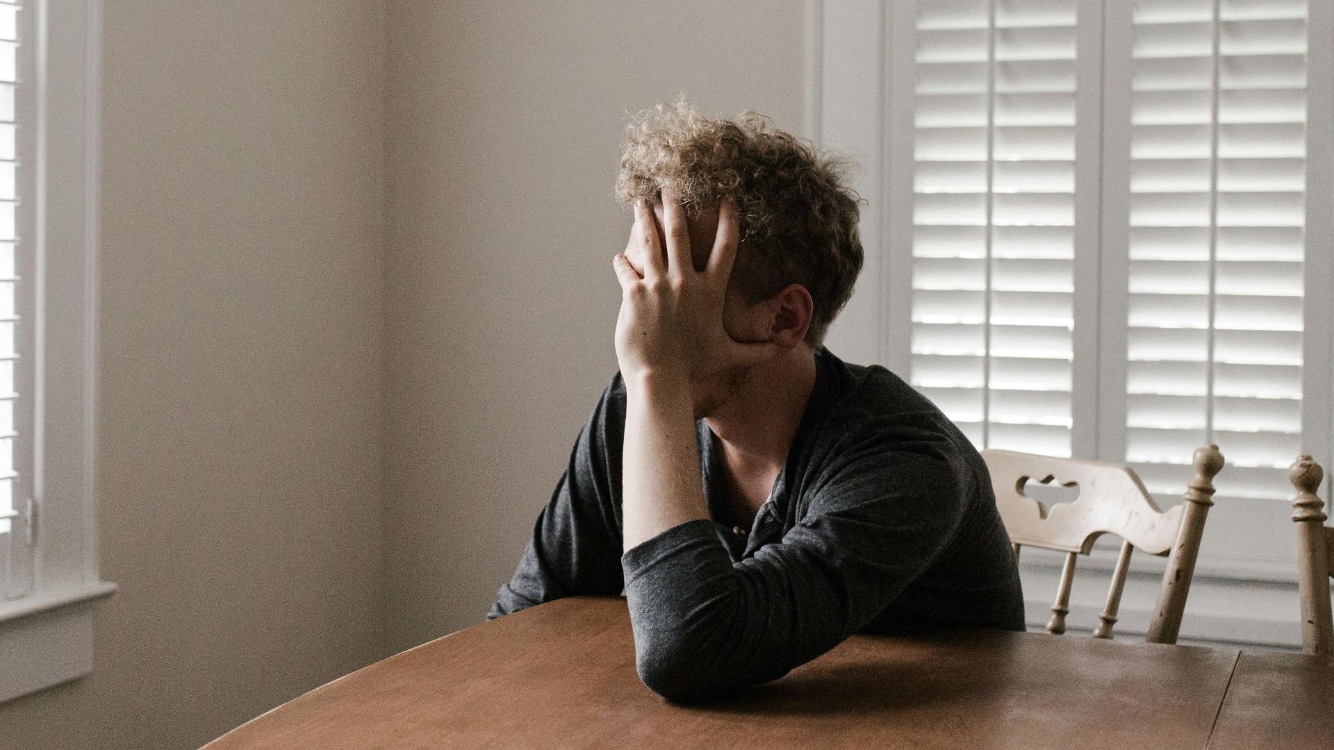 A man sits alone at a table in a bright room, displaying deep contemplation.