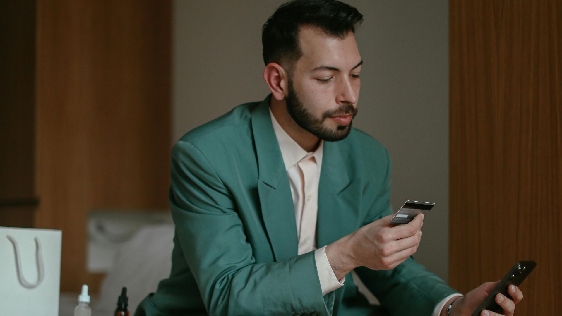 Man in casual business attire checks phone and bank card in a modern hotel room.
