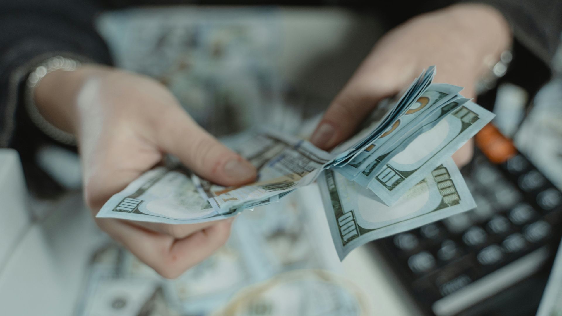 Close-up of hands counting hundred dollar bills with a calculator in the background.