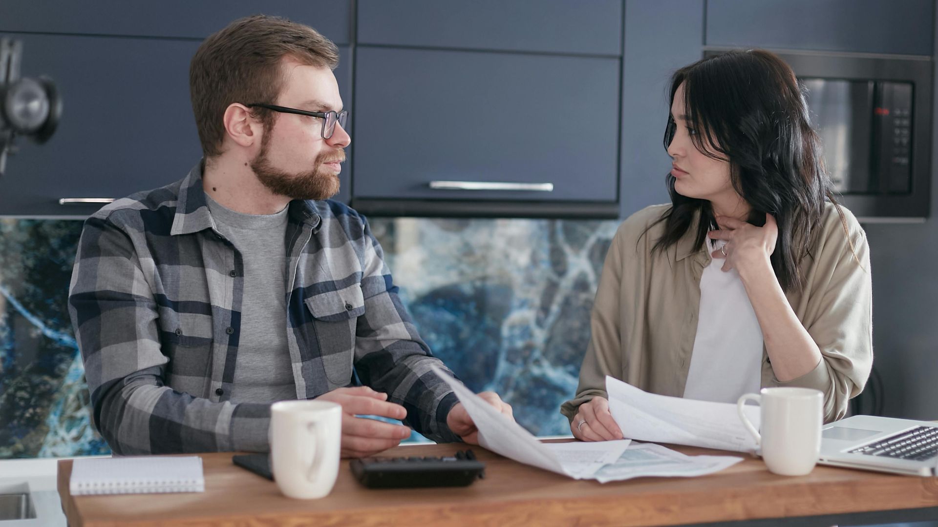 A young couple sitting at a table discussing bills and financial plans in a modern kitchen.