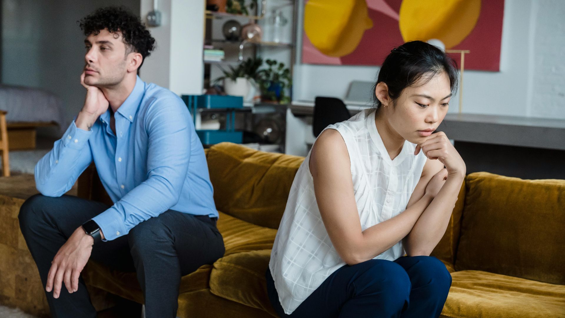 A couple sits indoors on a sofa, both appearing thoughtful and contemplative.