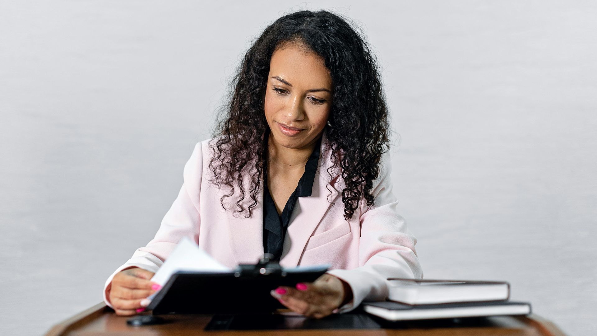 Confident woman with curly hair in a formal attire reviewing documents at a desk.