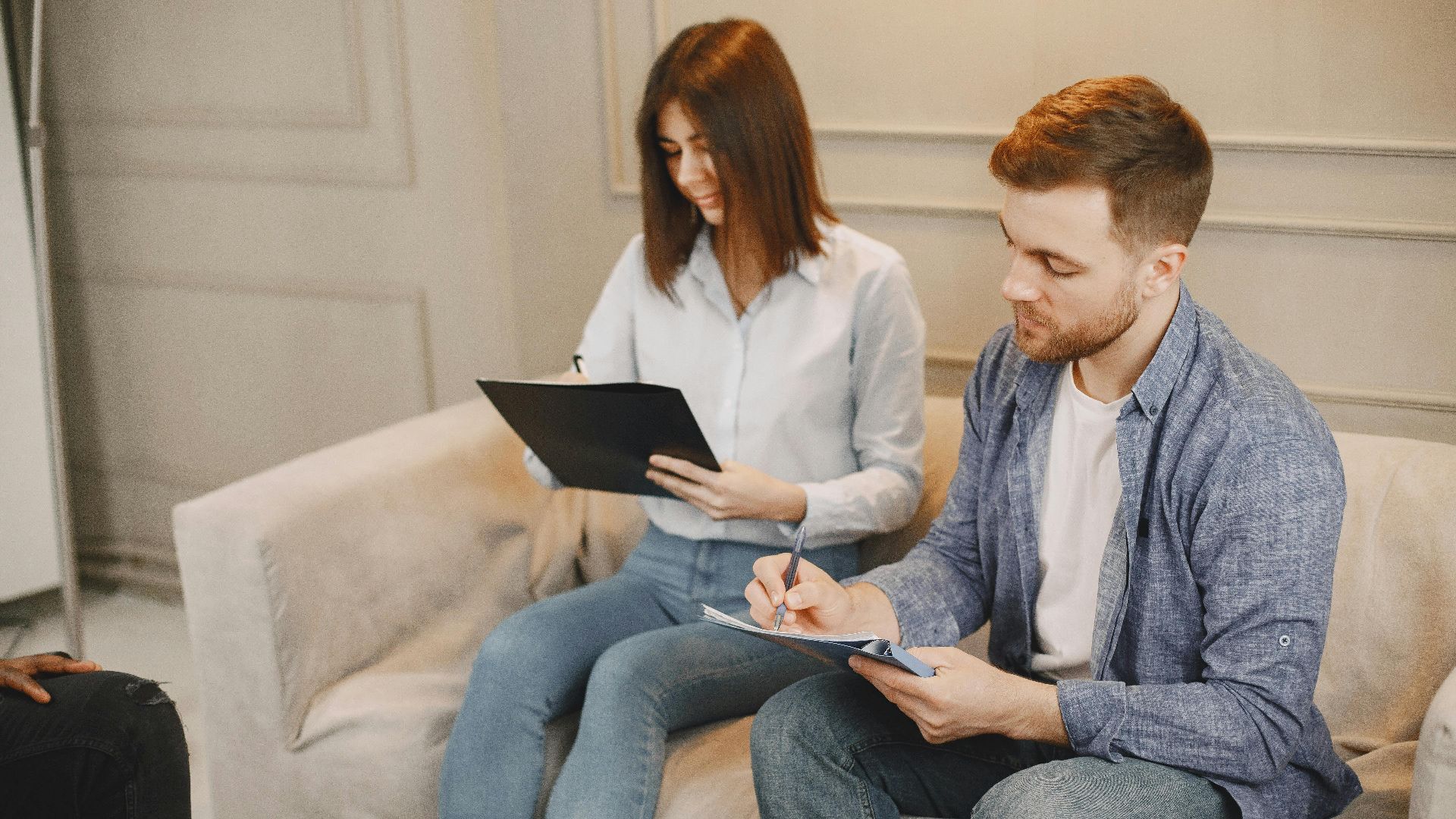 A young couple sitting on a sofa, writing and examining documents in a cozy indoor setting.