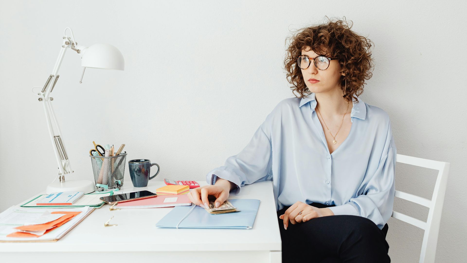 Woman in blue shirt with glasses sits at white desk with documents and phone.