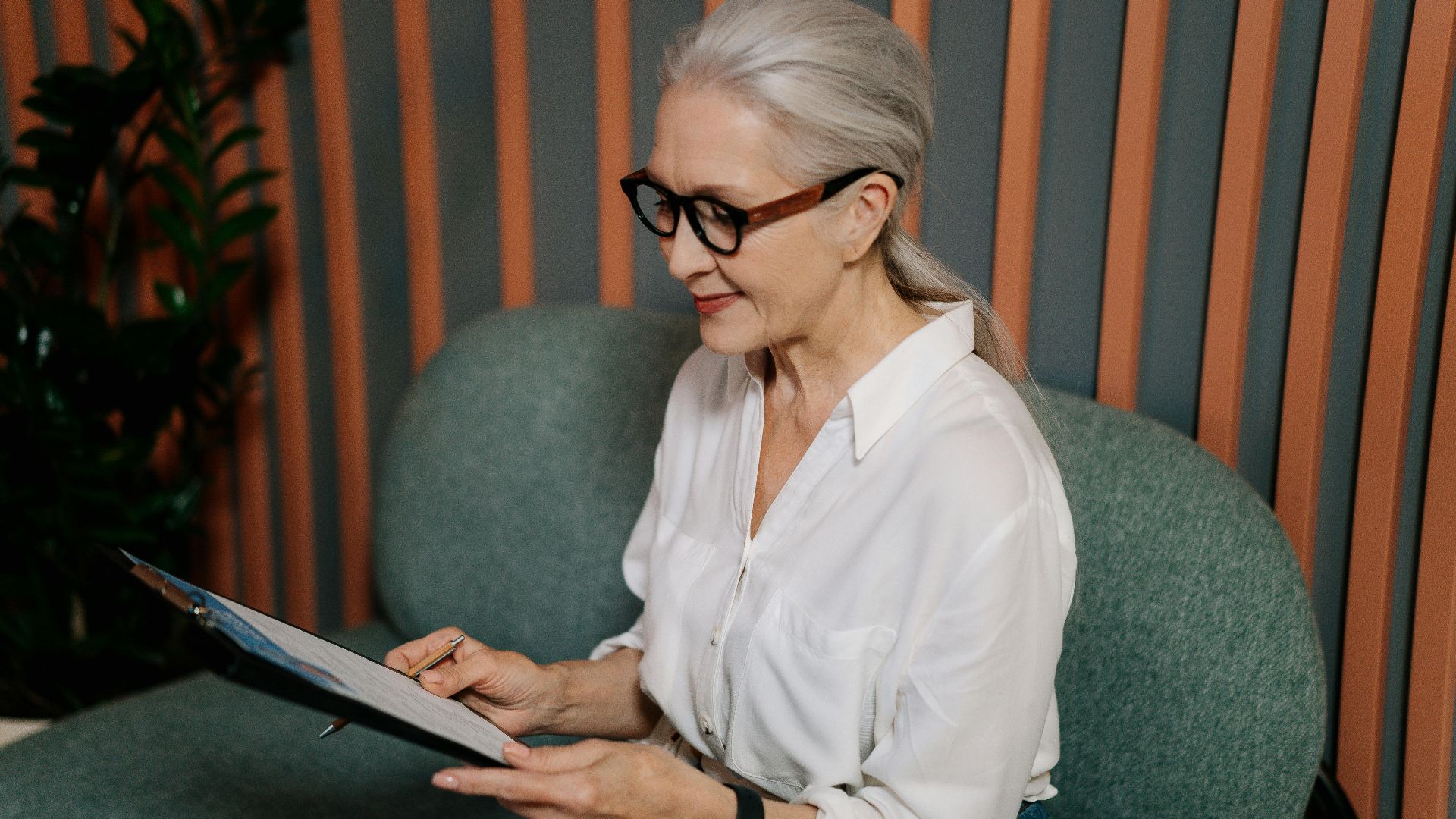 Elderly woman in glasses reviewing documents on clipboard, sitting indoors with a focused expression.