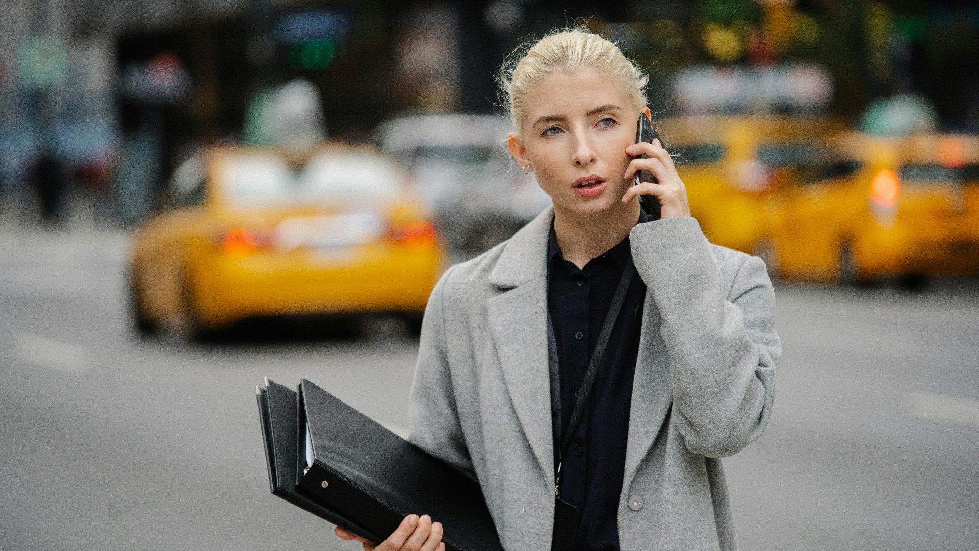 Focused young businesswoman in gray coat standing on busy street with folders and having conversation on mobile phone