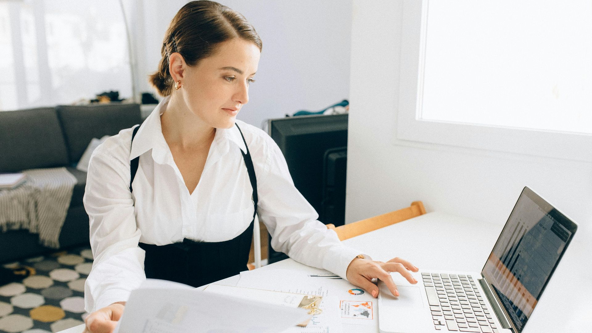 Woman at desk reviewing business reports on laptop, holding papers in a modern office setting.