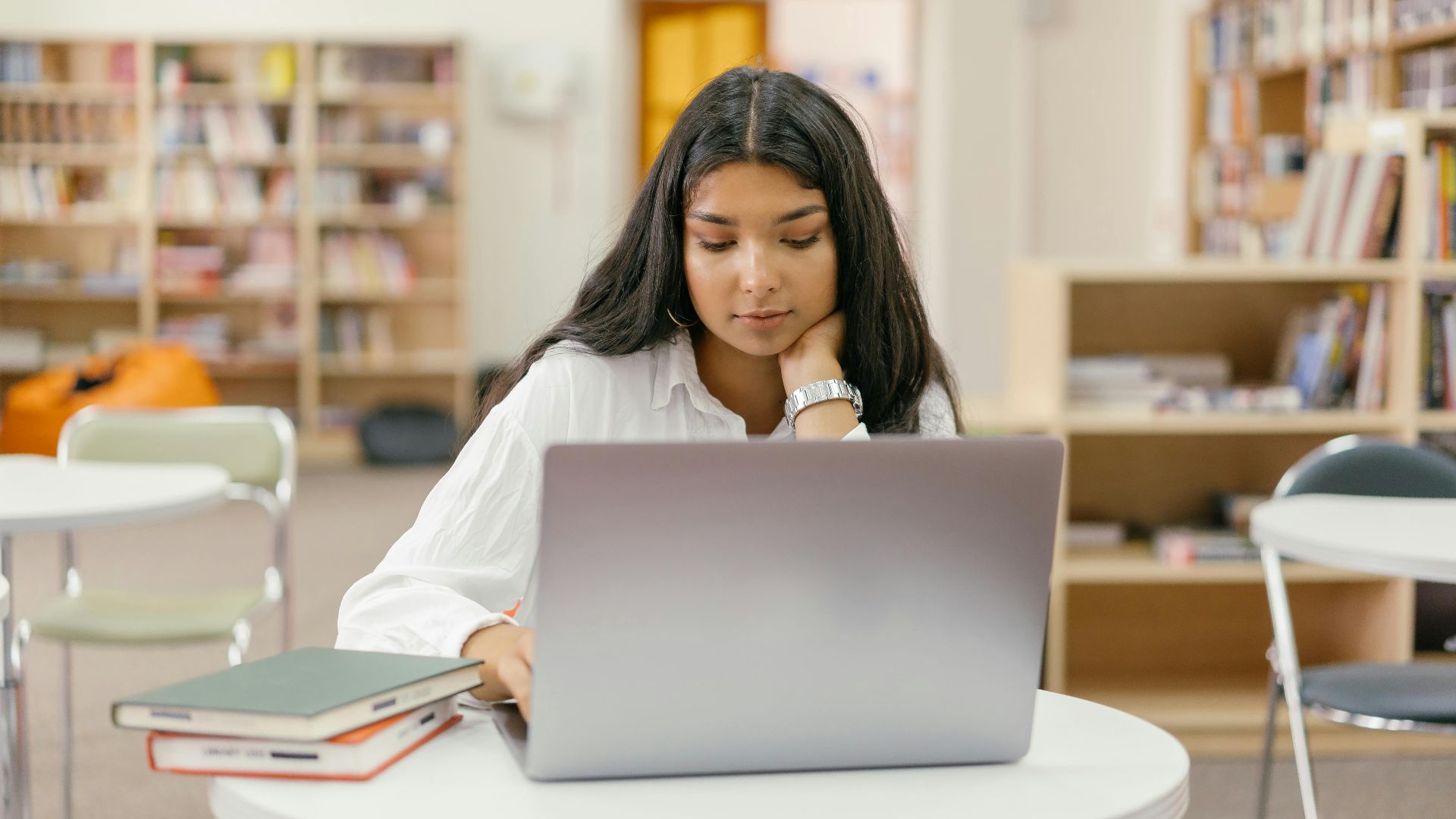 A young woman in a library studying online with a laptop and books around her.