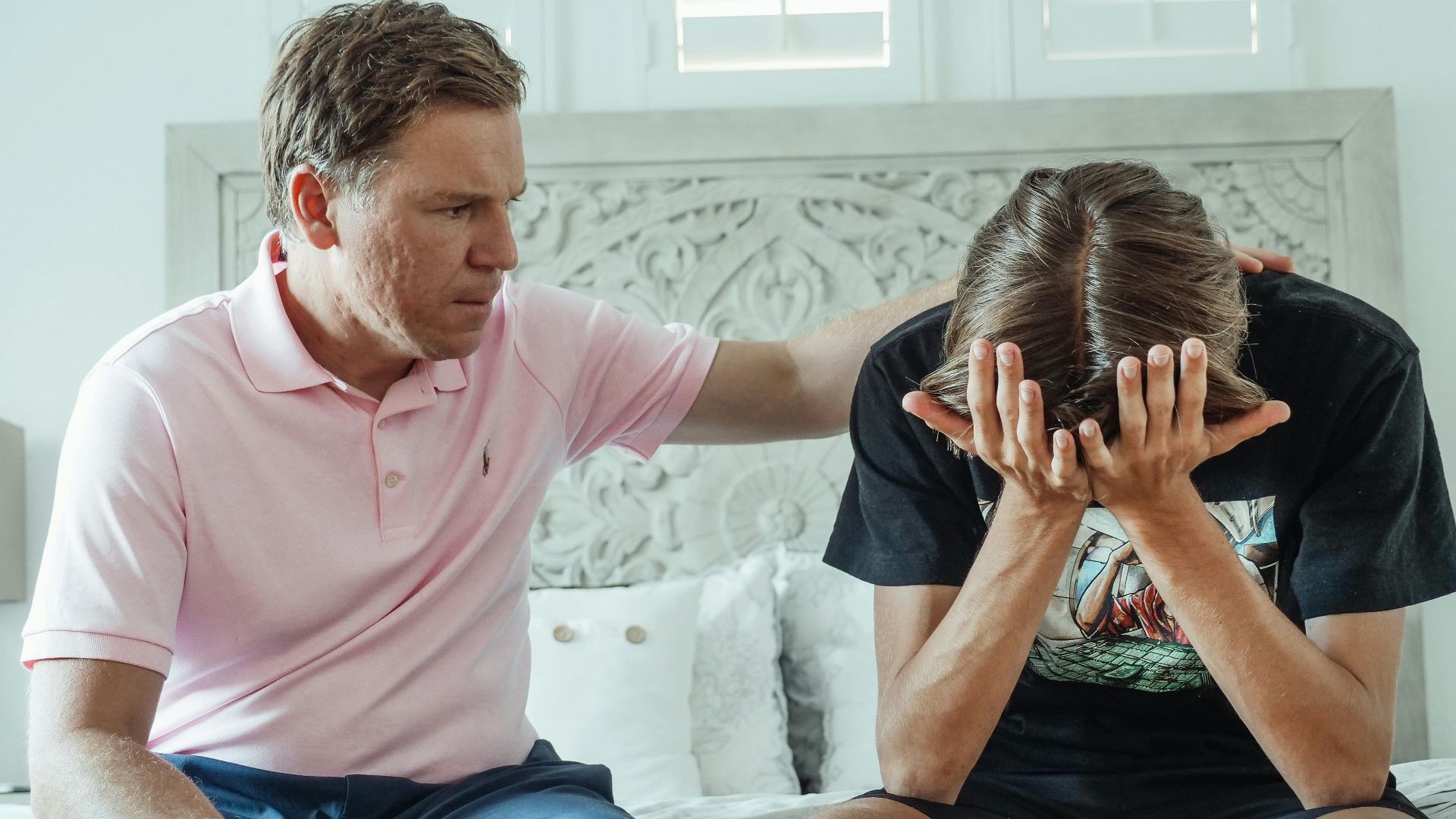 A compassionate father consoles his upset teenage son on a bed indoors.
