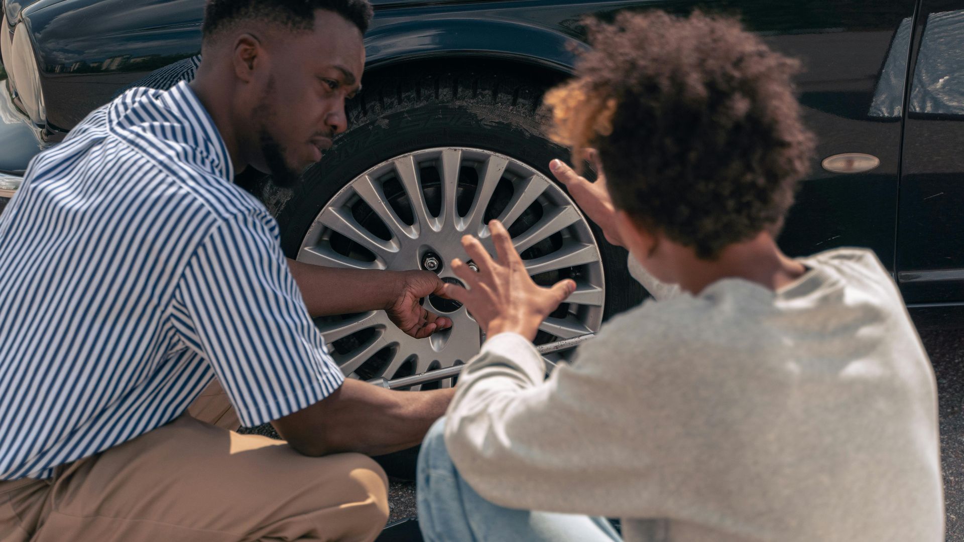 A father instructs his teenage son on how to change a car tire outdoors.