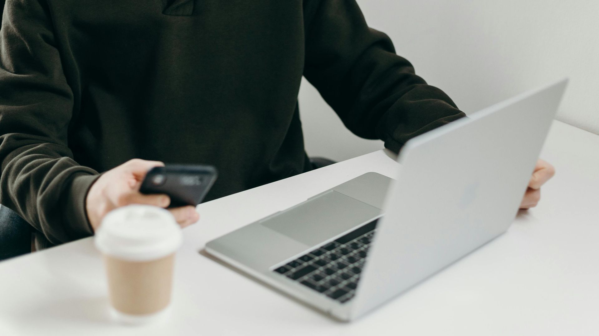 Casually dressed man working from home using a laptop and smartphone, with a coffee cup nearby.