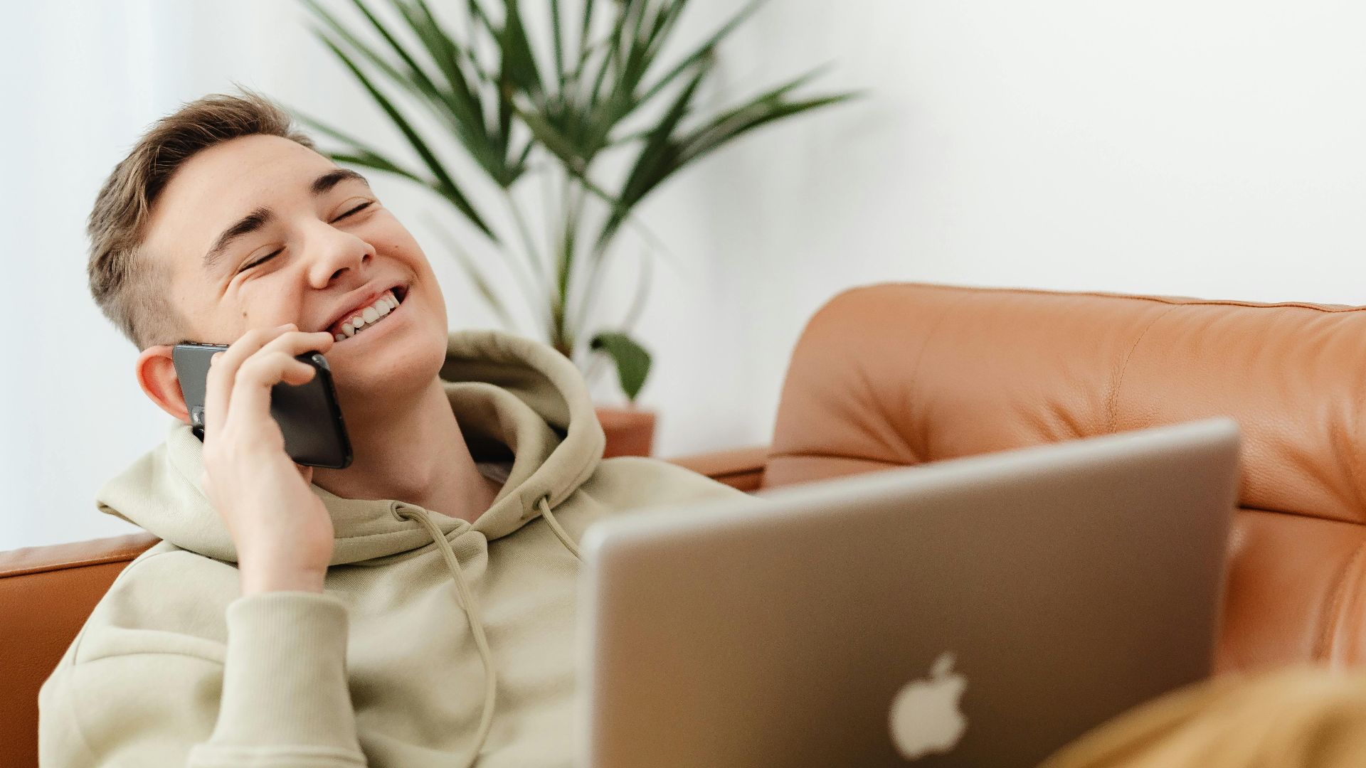 Smiling young man talking on cellphone, using laptop on sofa at home.