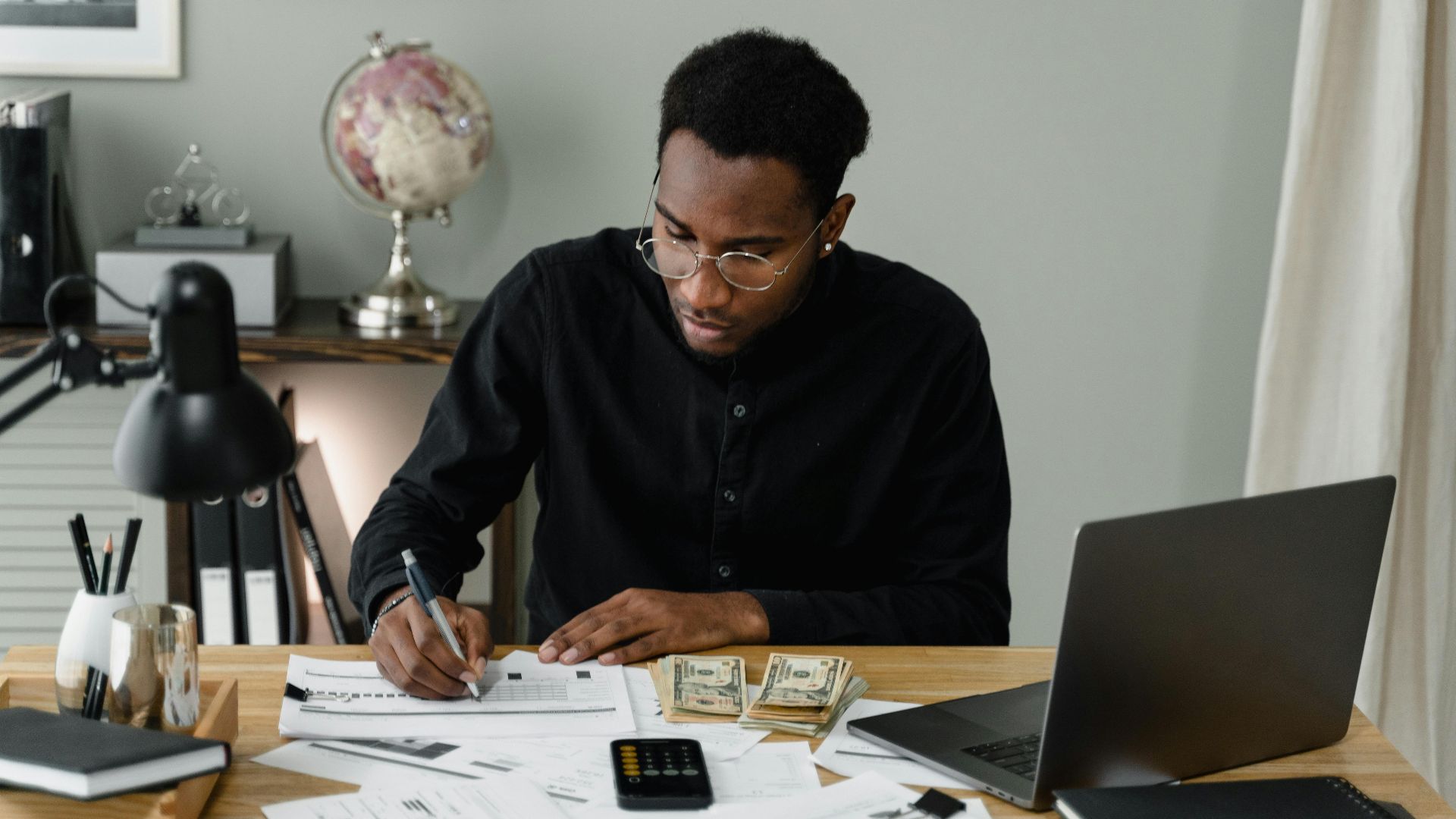 An adult man calculates expenses, using a laptop and documents at a desk in a home office setting.