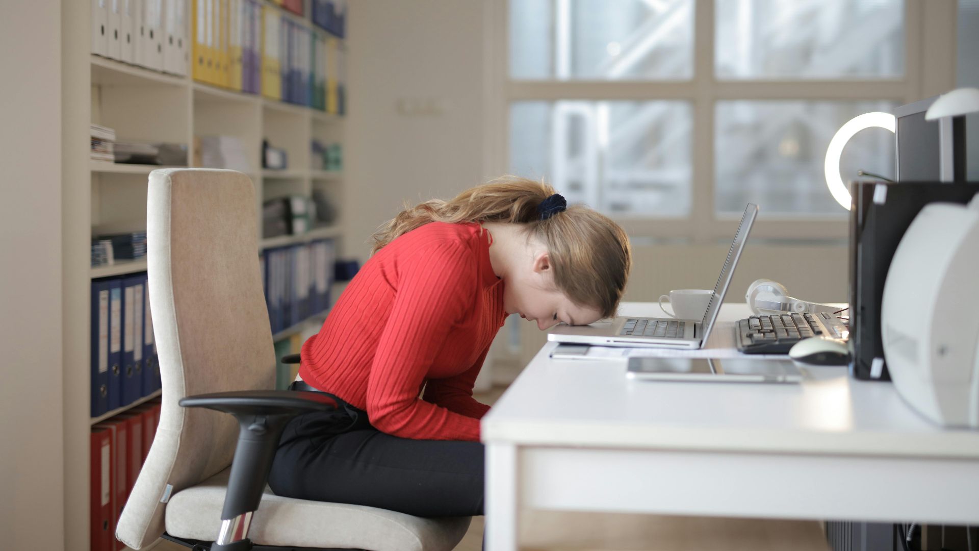 Tired woman in red sweater naps on office desk beside laptop, overwhelmed by remote work.