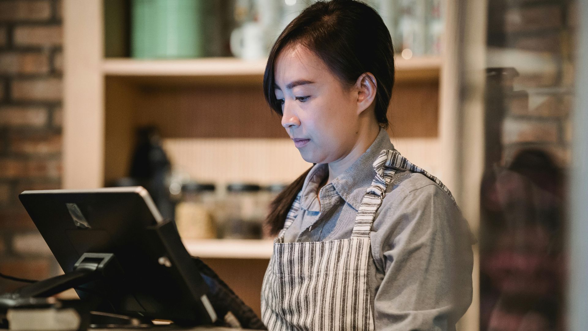 A barista in a striped apron working diligently at a cafe counter, using a point-of-sale system.