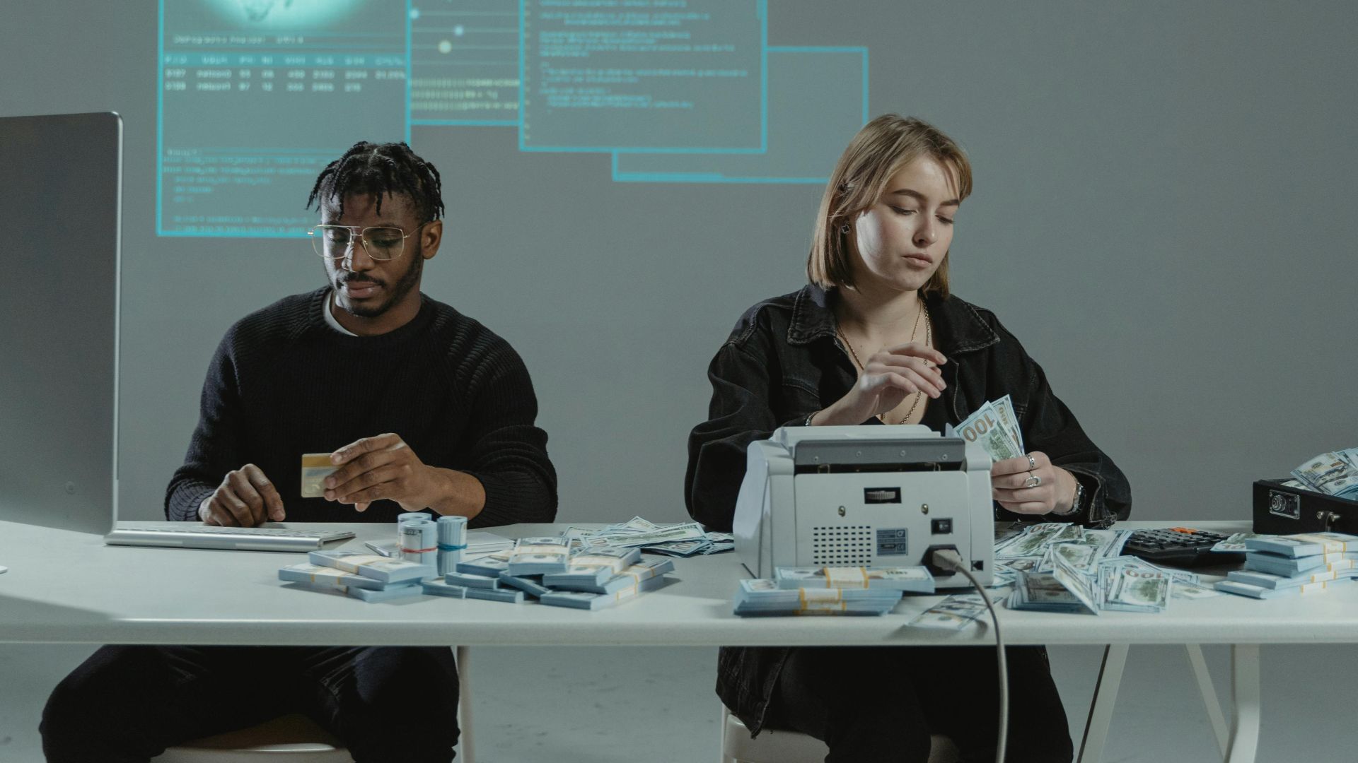Two adults counting money in an office with a tech-themed background.