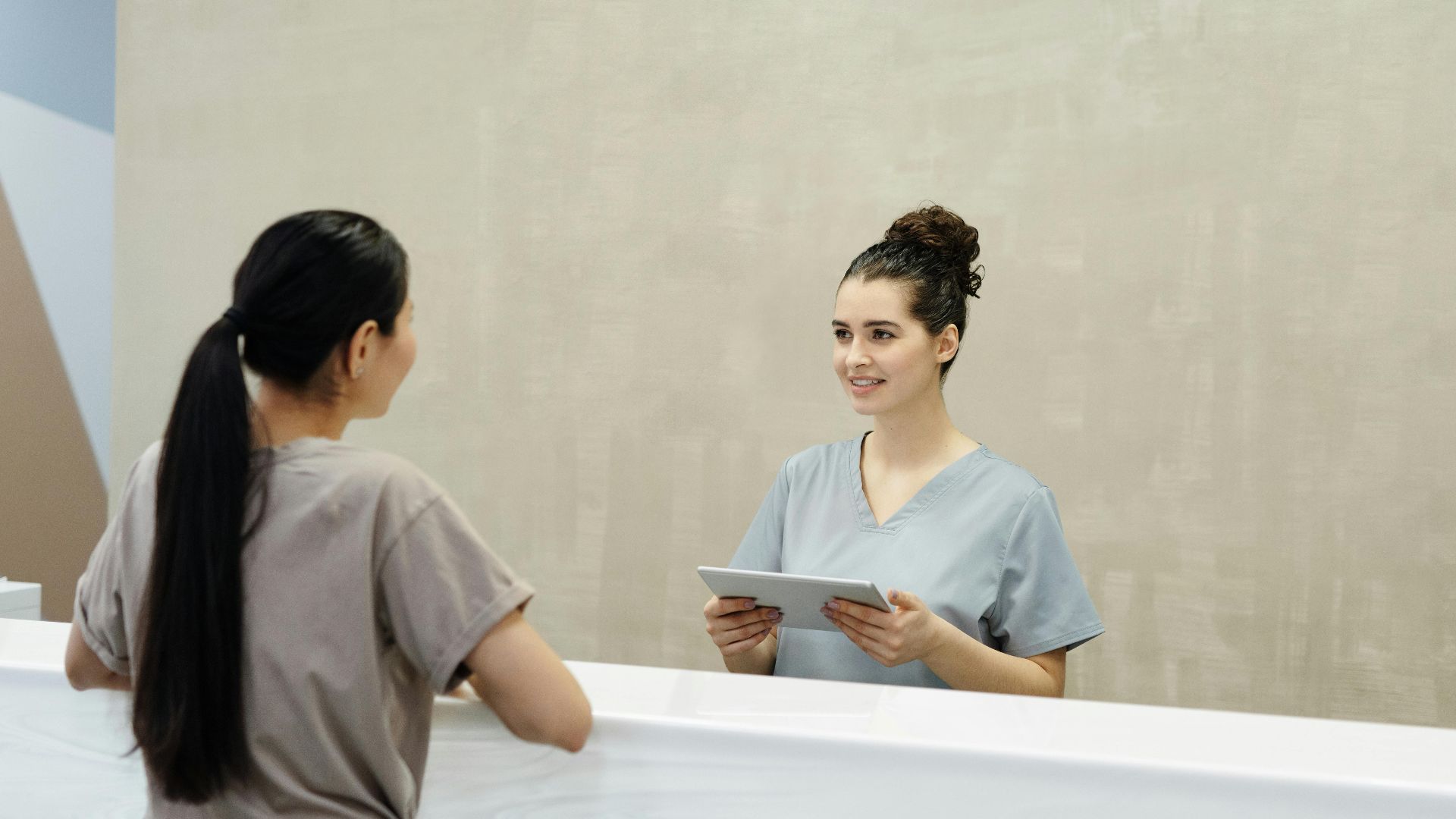 A smiling receptionist with a tablet assists a patient at a medical facility reception desk.