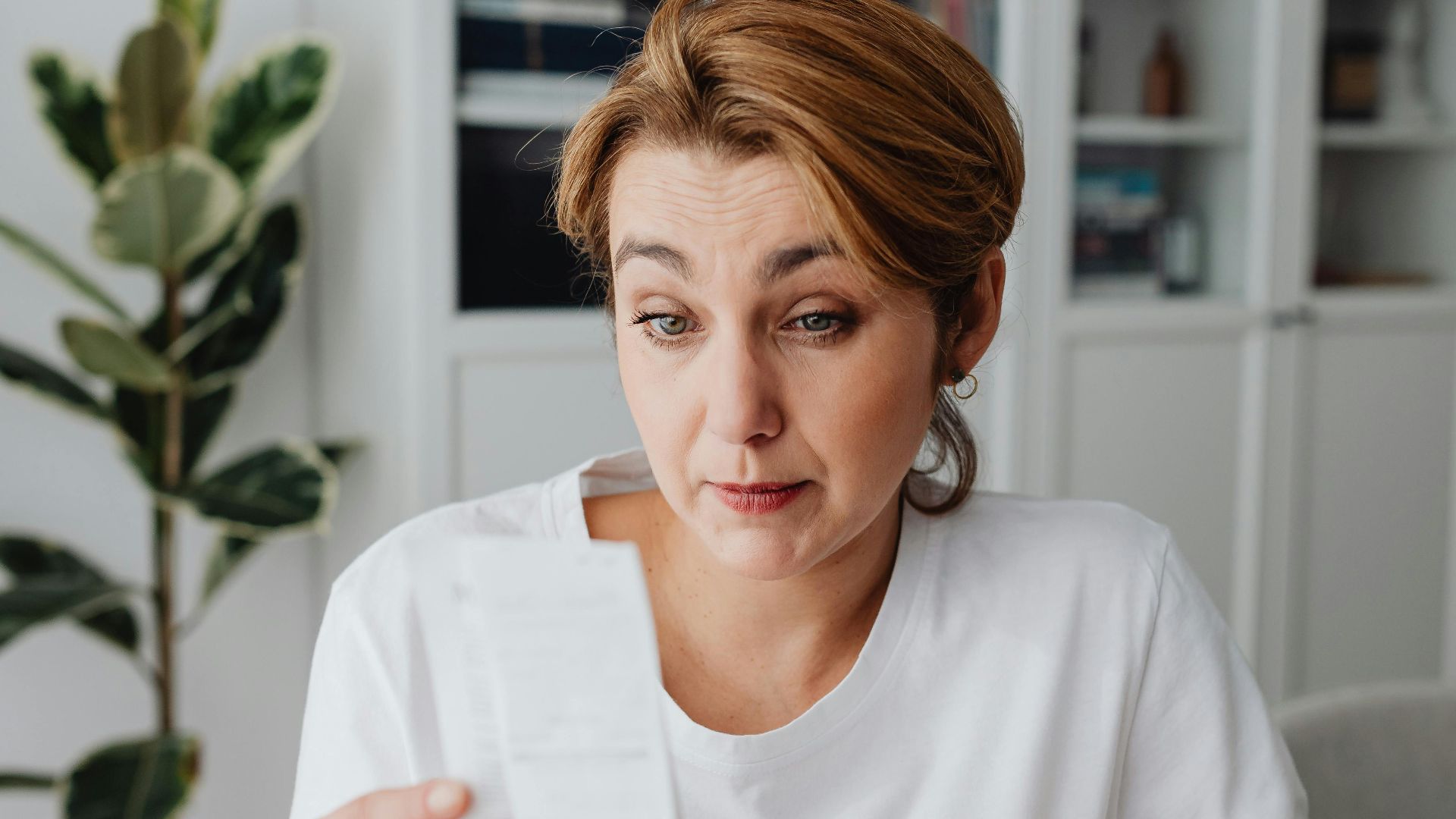 An adult woman with a surprised expression looks at a receipt at her desk.