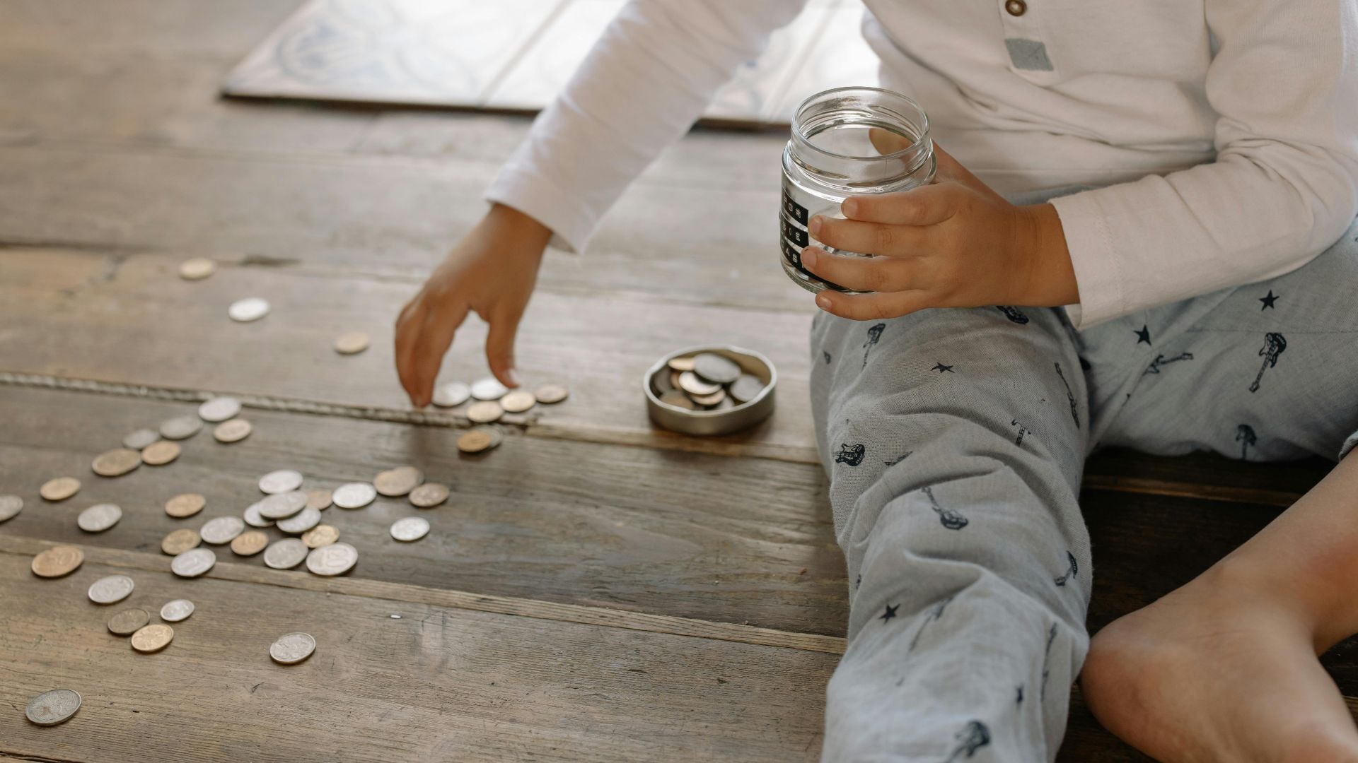 A young child collects coins into a jar on a wooden floor, symbolizing savings.
