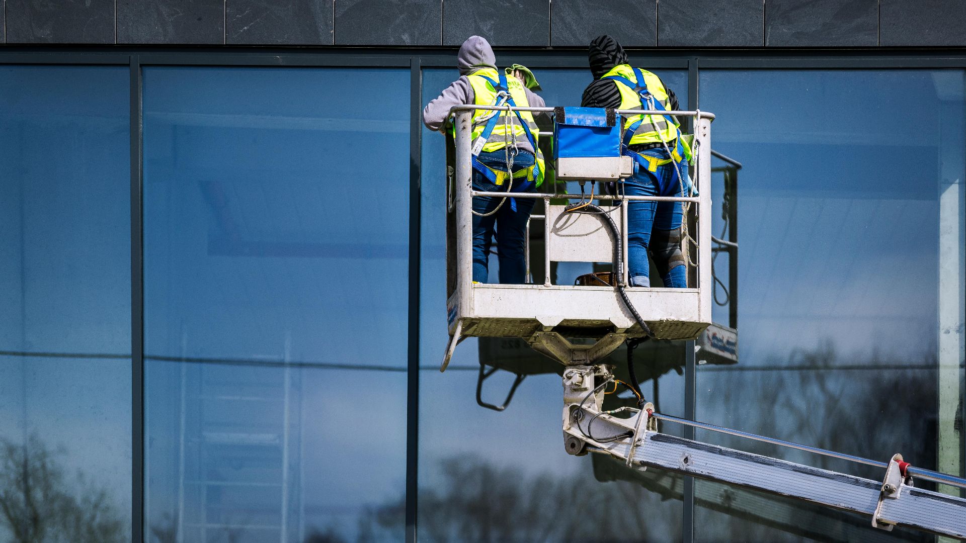 Two men on a hydraulic lift cleaning windows of a modern skyscraper. Urban reflection visible in the glass.