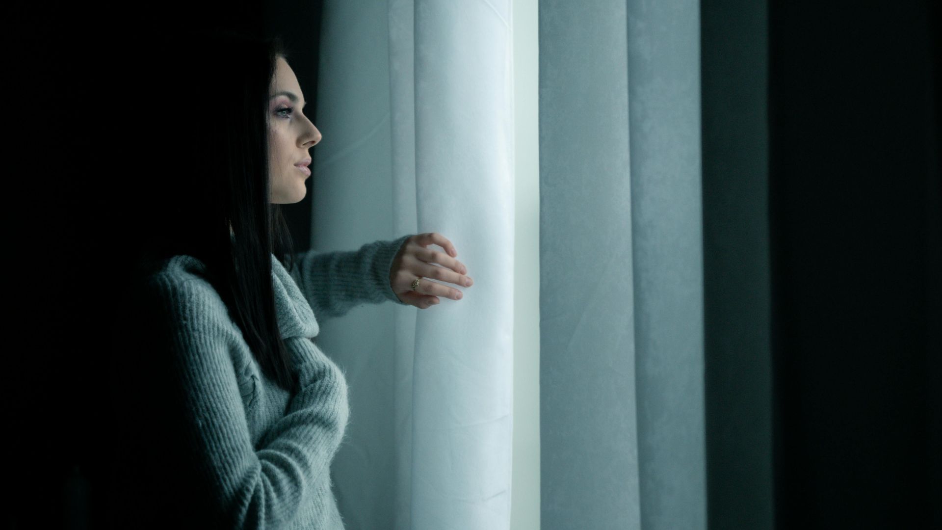 Portrait of a woman looking out a window with a thoughtful expression in a dimly lit room.