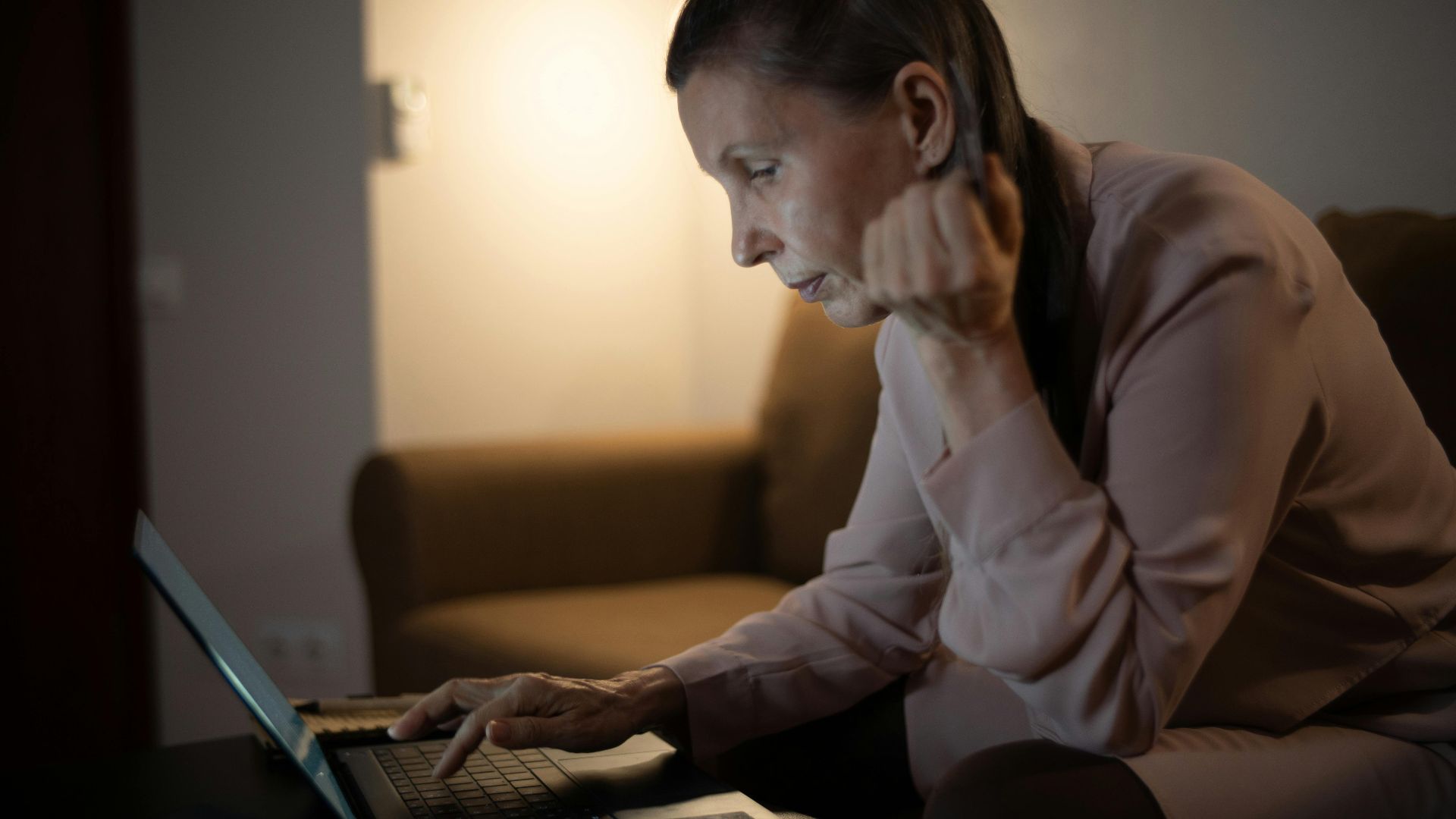 Senior woman using laptop in a dimly lit living room, focused on screen.