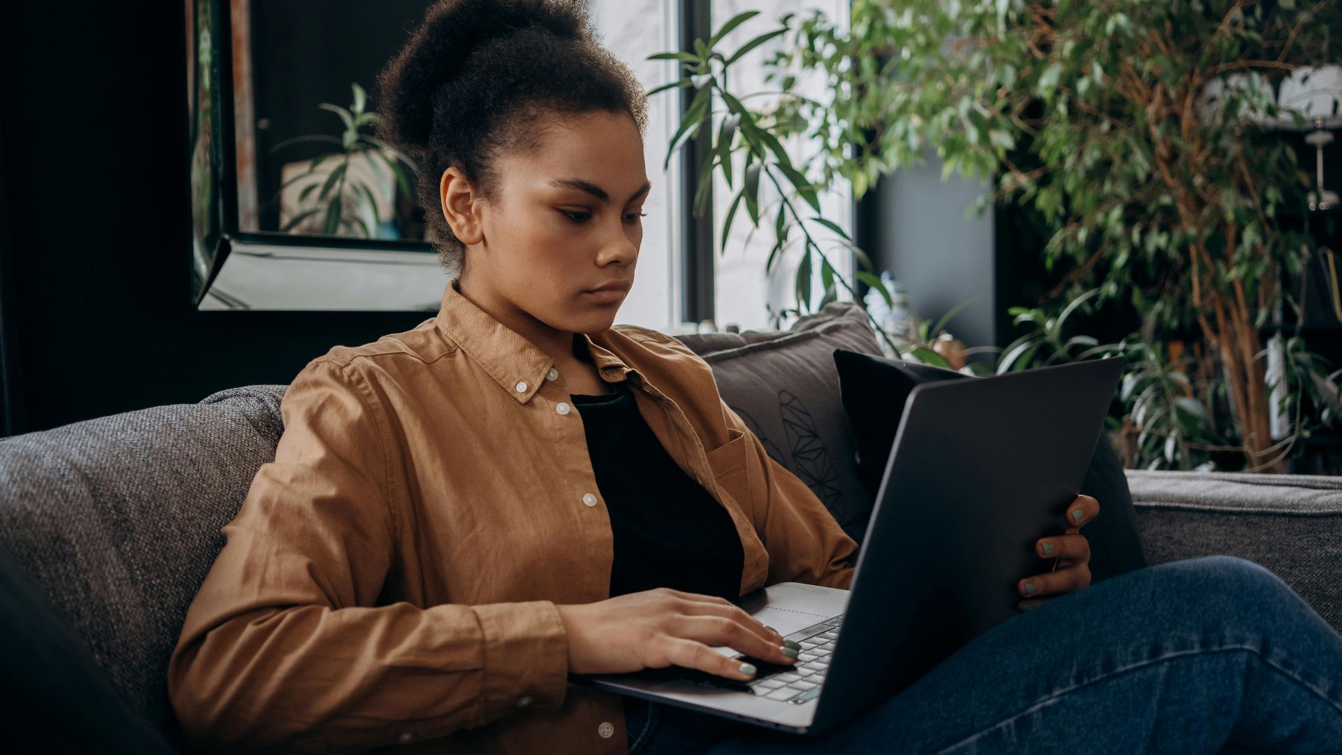 Focused young woman using a laptop on the couch, working remotely indoors.