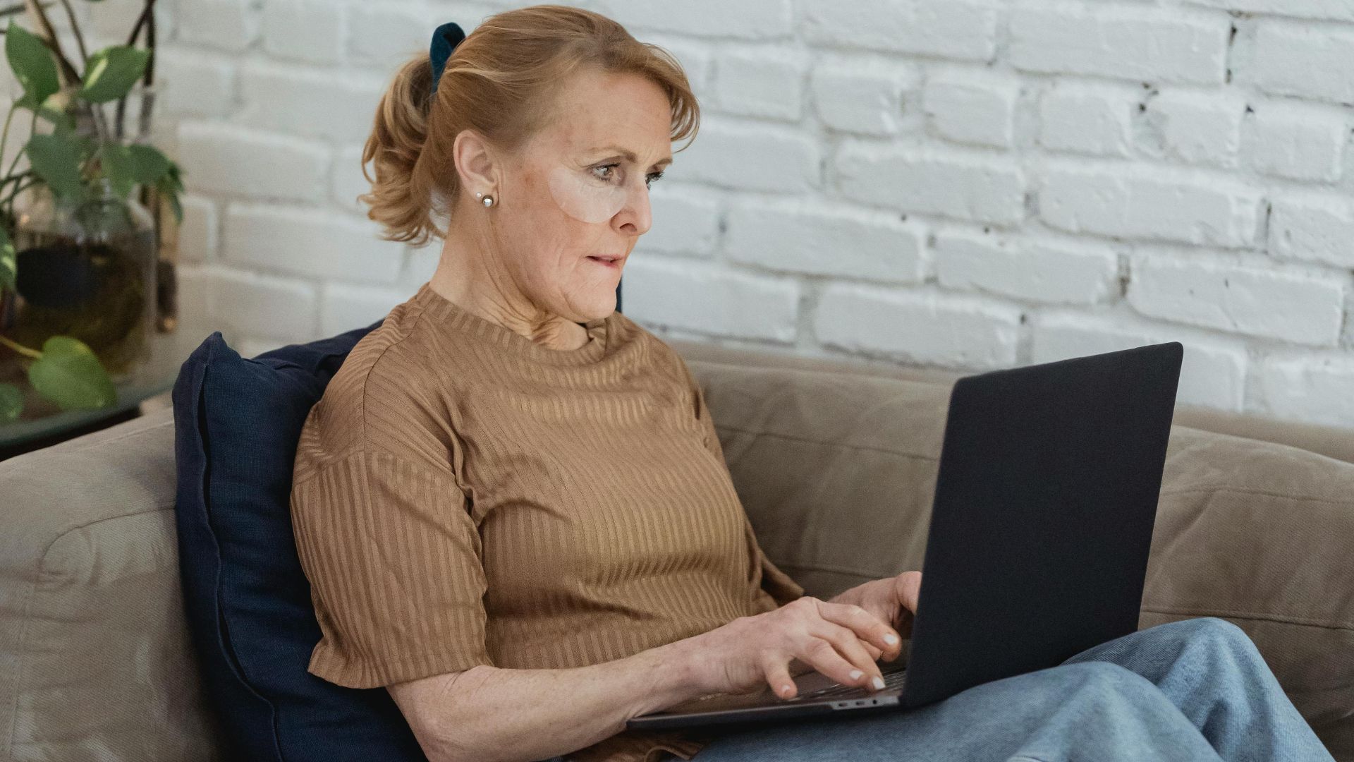 Senior woman comfortably working on her laptop indoors, surrounded by plants and cozy home decor.