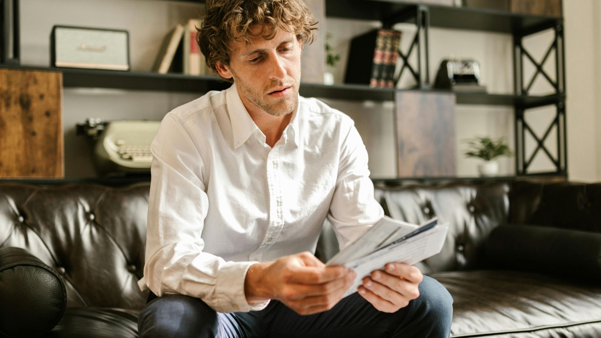 Adult man sitting on leather sofa in home office, reading documents.