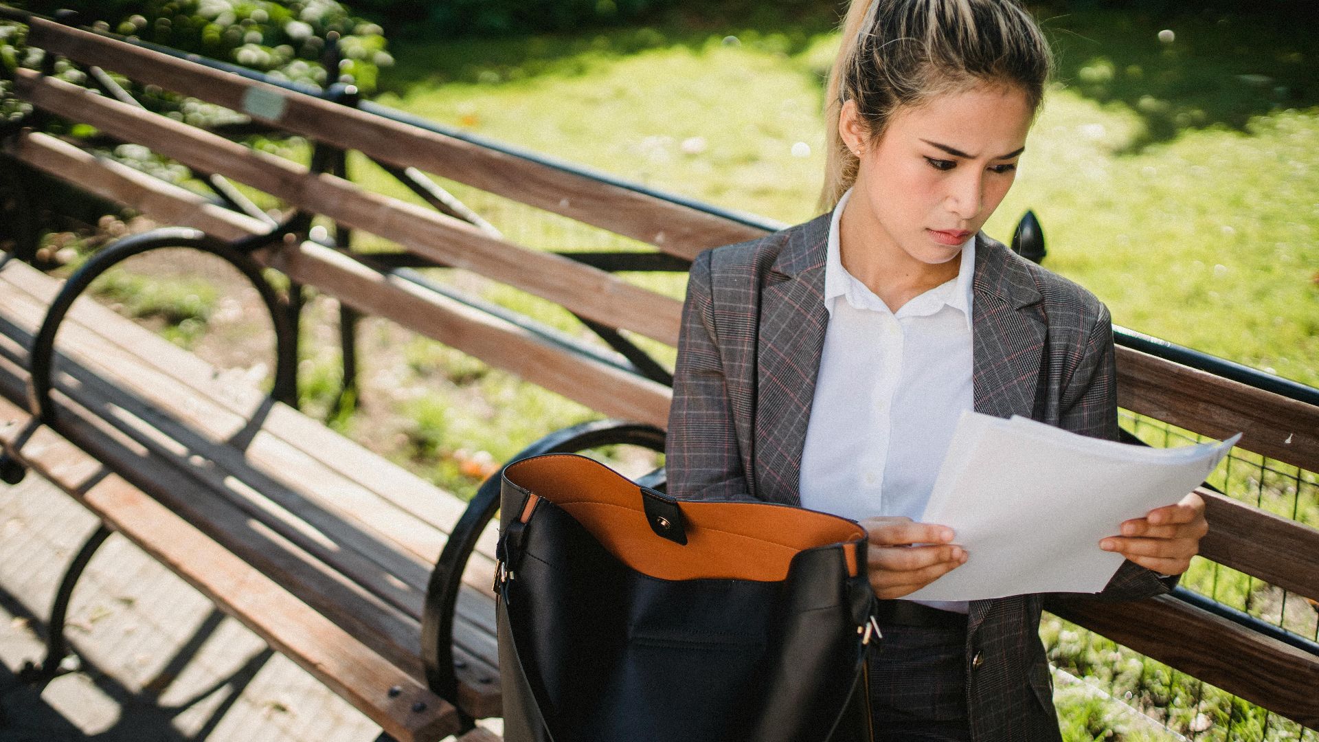 Professional woman in suit reading documents on park bench, showcasing business elegance outdoors.