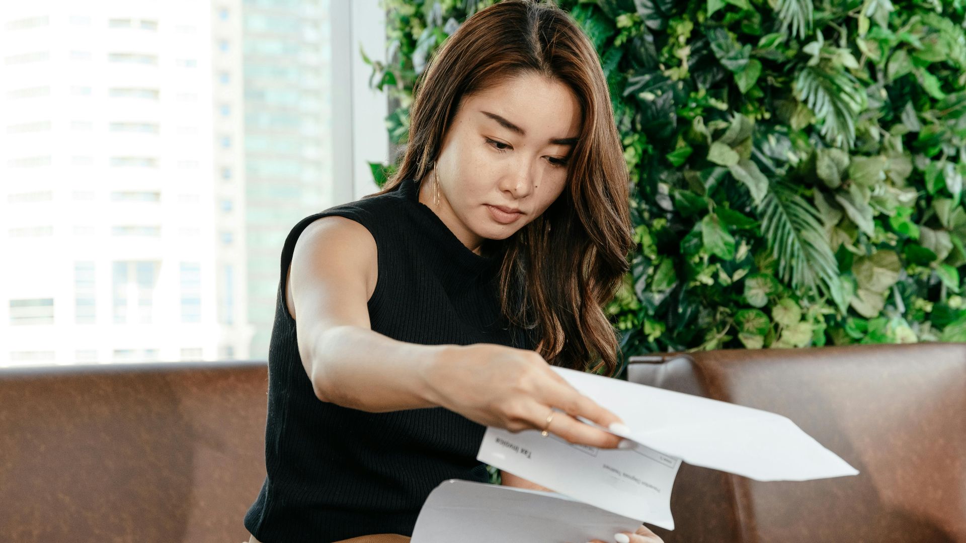 Focused Asian female turning pages of document while sitting on sofa during paperwork in modern workspace with green deciduous plant