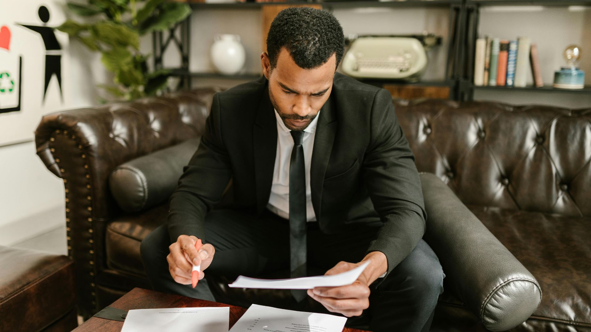 A businessman in a suit attentively reviews paperwork on a brown leather sofa.