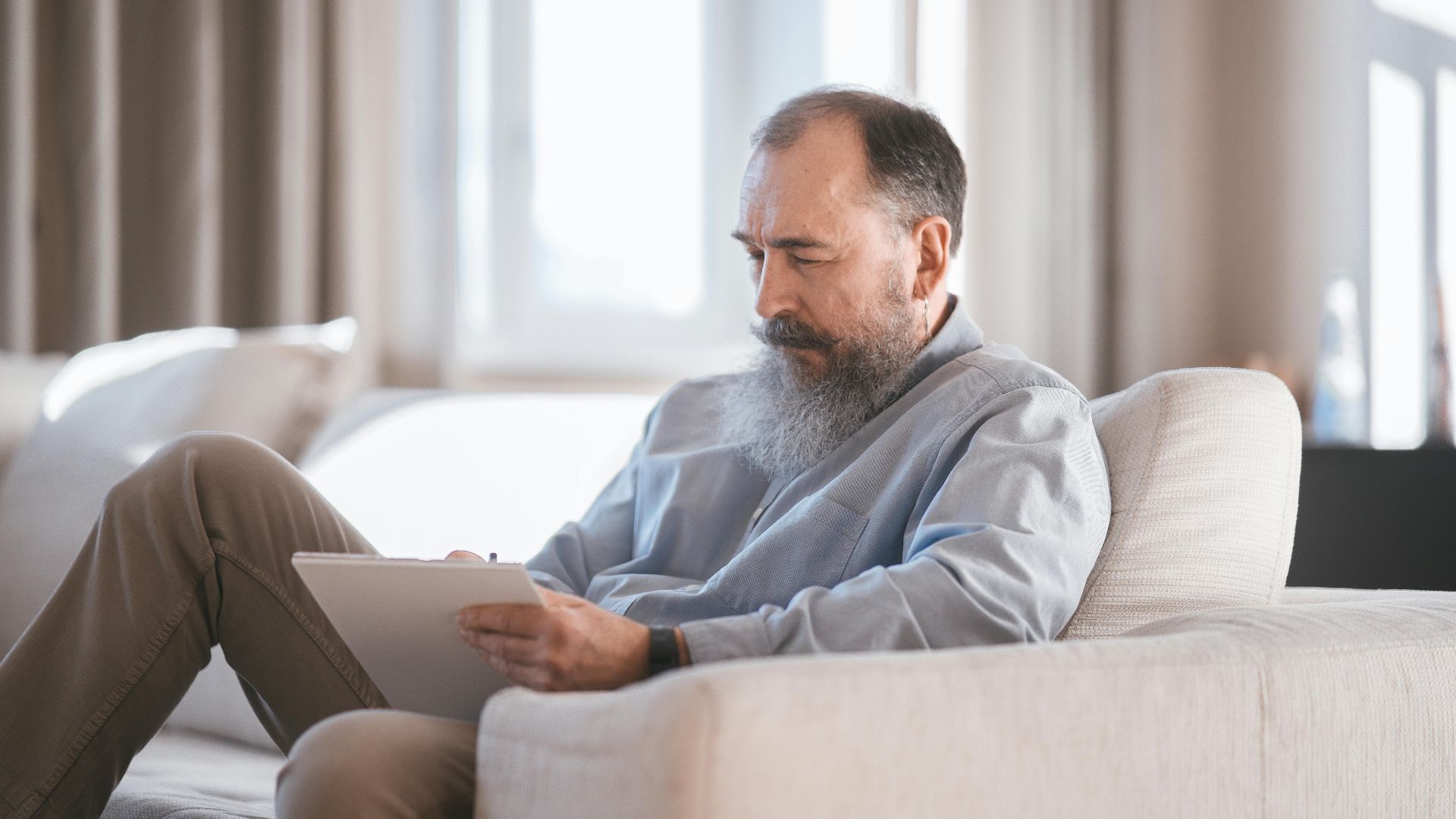 Elderly man sitting on sofa writing on a tablet with focus and concentration.