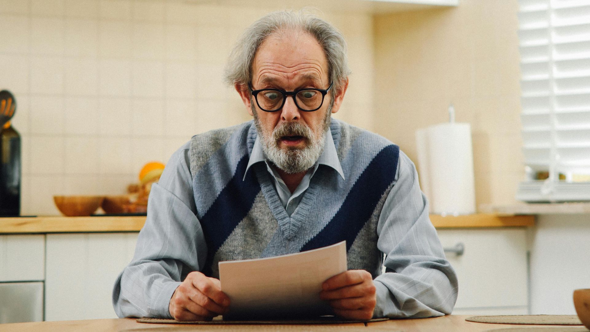 Senior man with eyeglasses reading a document at kitchen table, reflecting surprise.