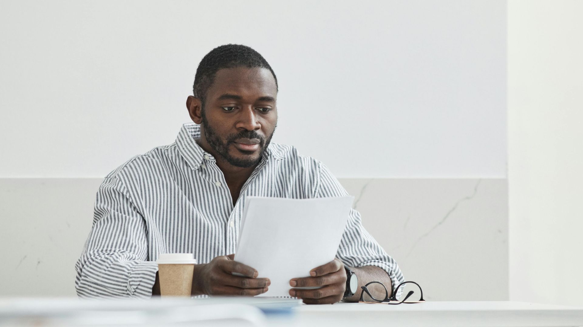 African American man sitting indoors, reading papers with a coffee cup nearby.
