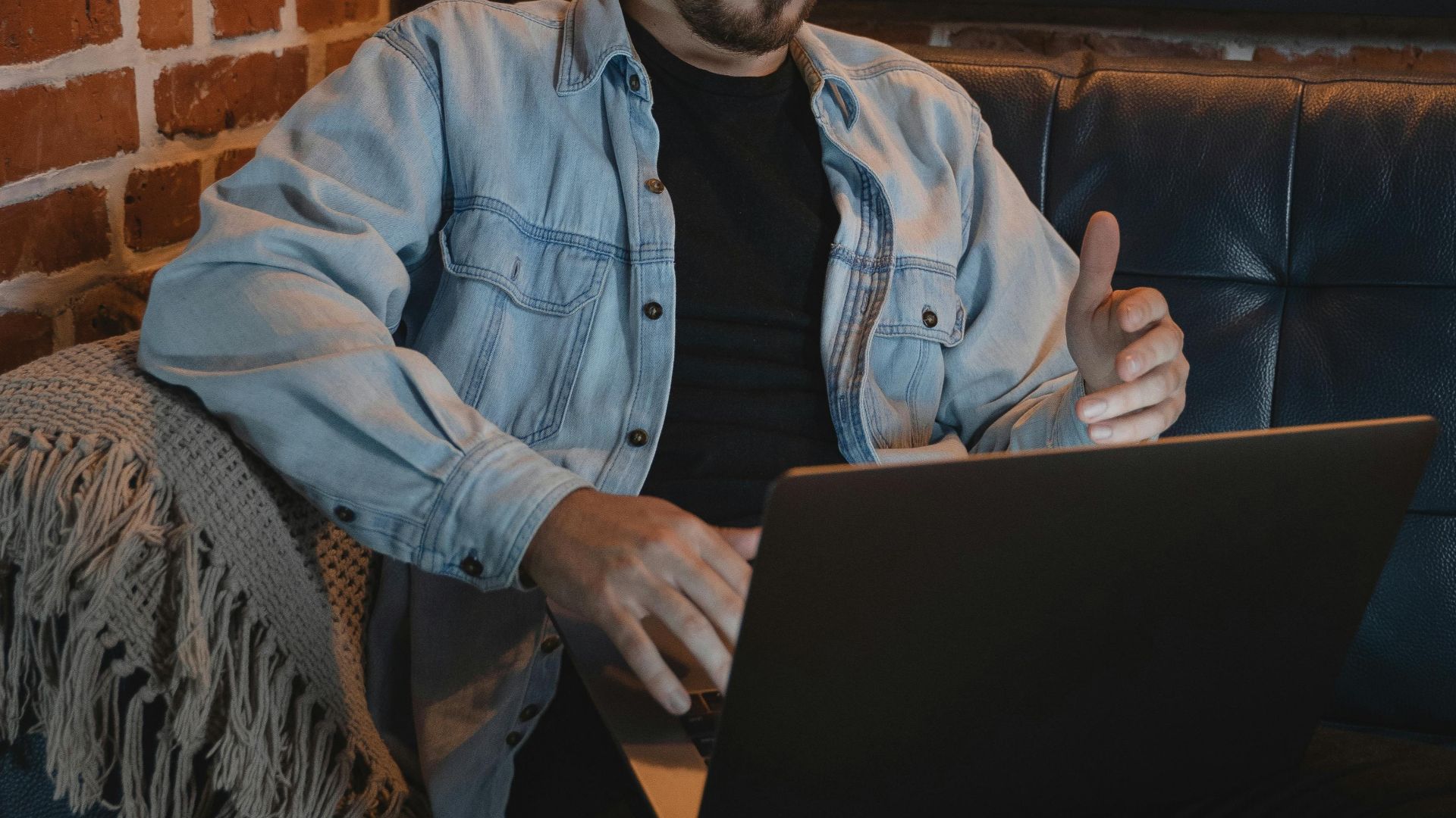 Adult man working remotely on a laptop in a cozy urban setting with exposed brick walls.