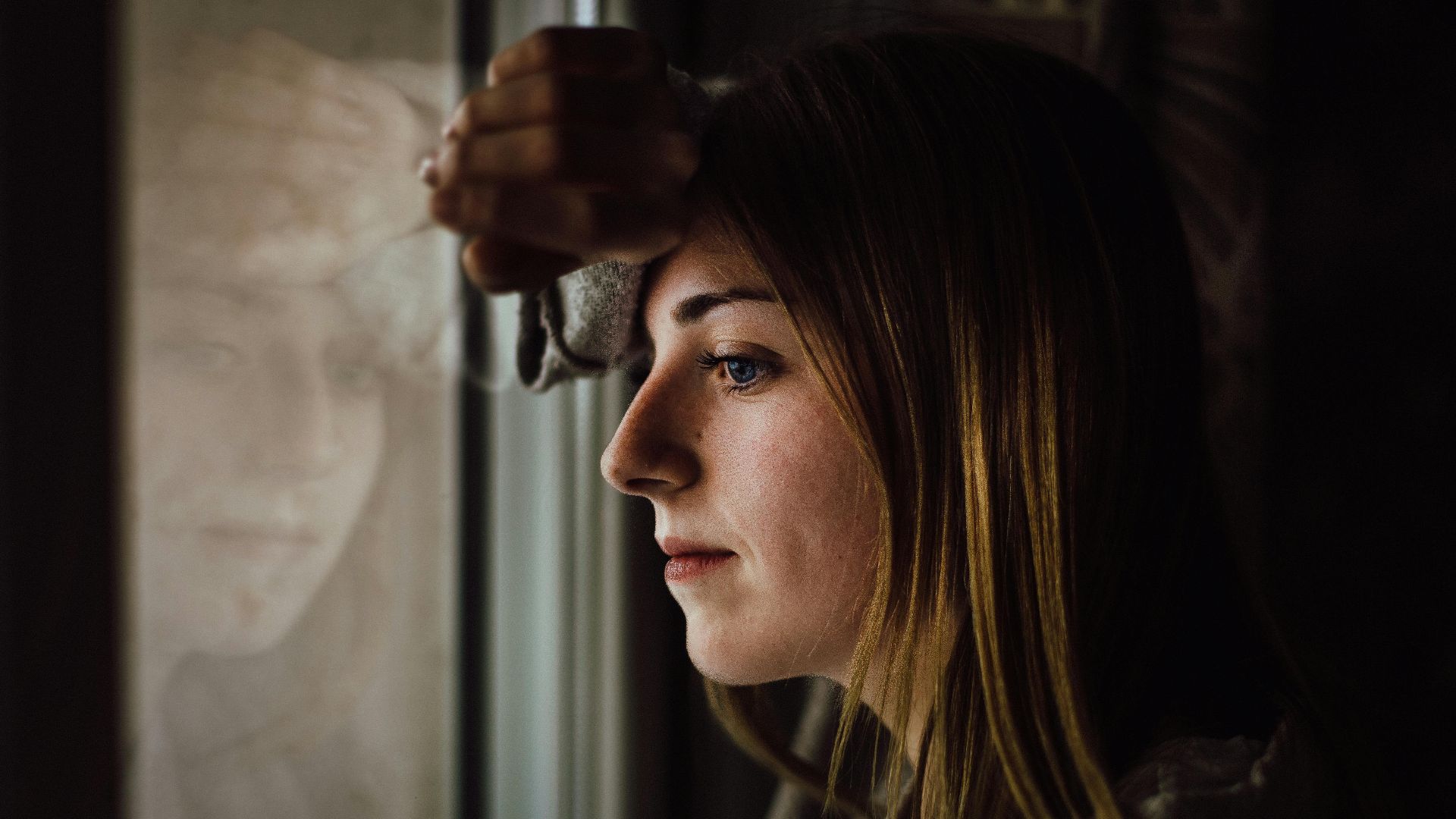 A contemplative young woman looks out a window with a reflective expression, indoors.