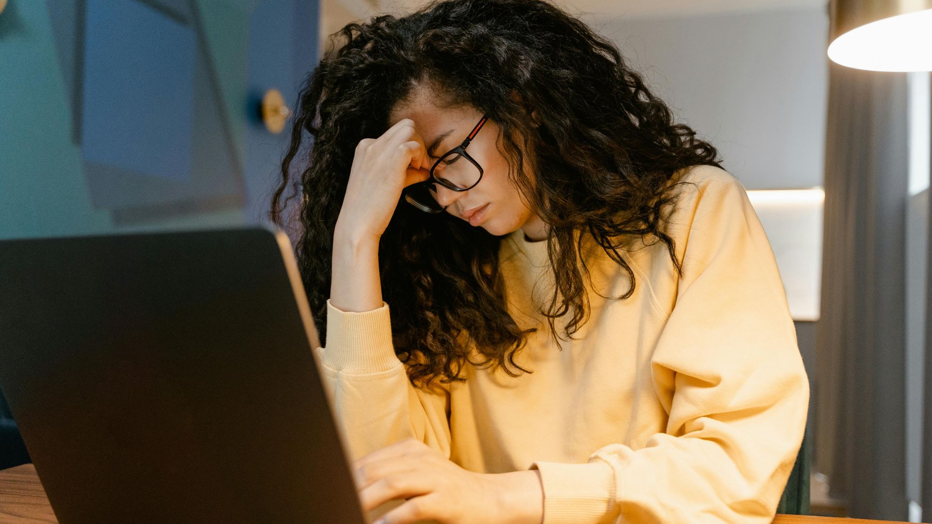 A tired woman with curly hair and glasses experiencing stress while working on a laptop.