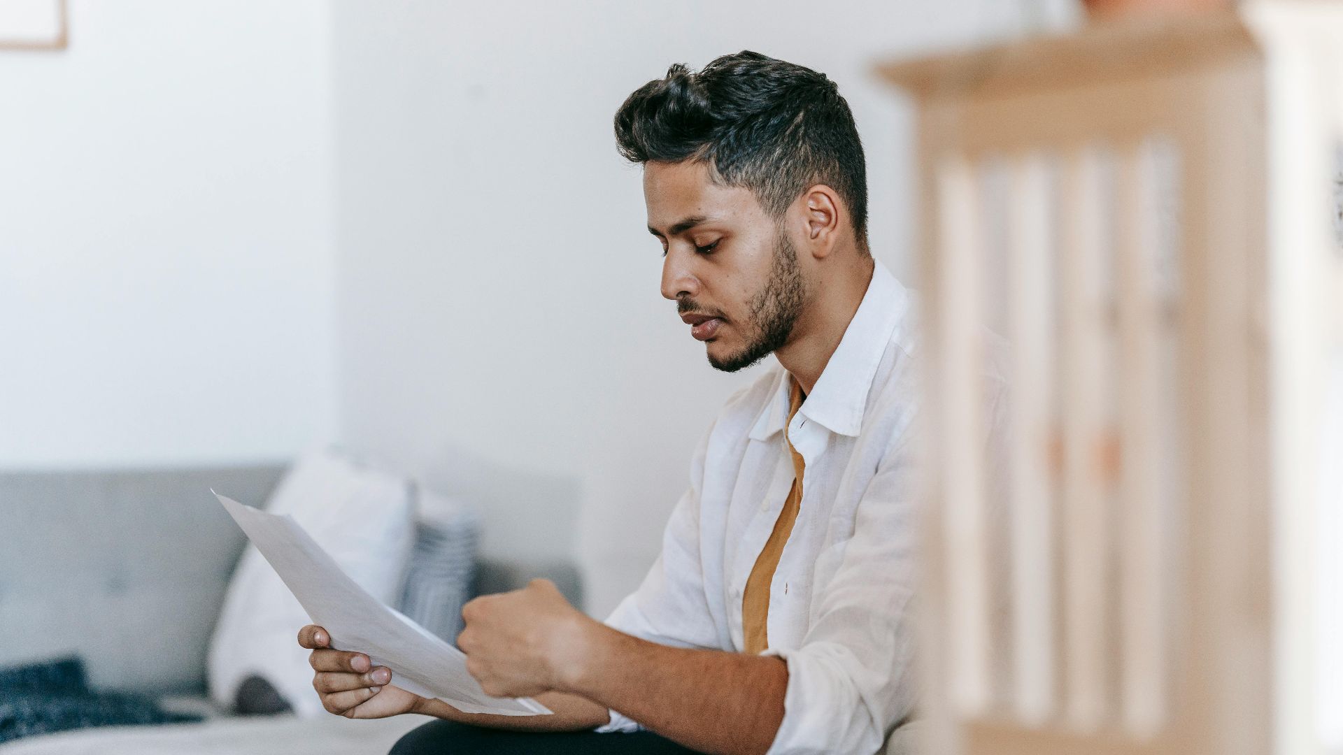 Young man in a white shirt reading papers at home, appearing thoughtful and focused.