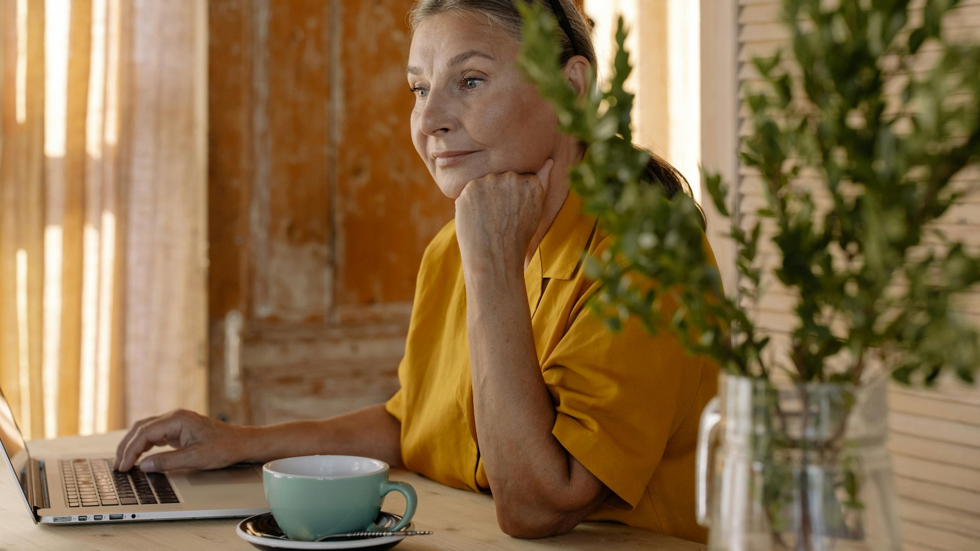 Elderly woman sitting at a table using a laptop, relaxing in a cozy home setting.